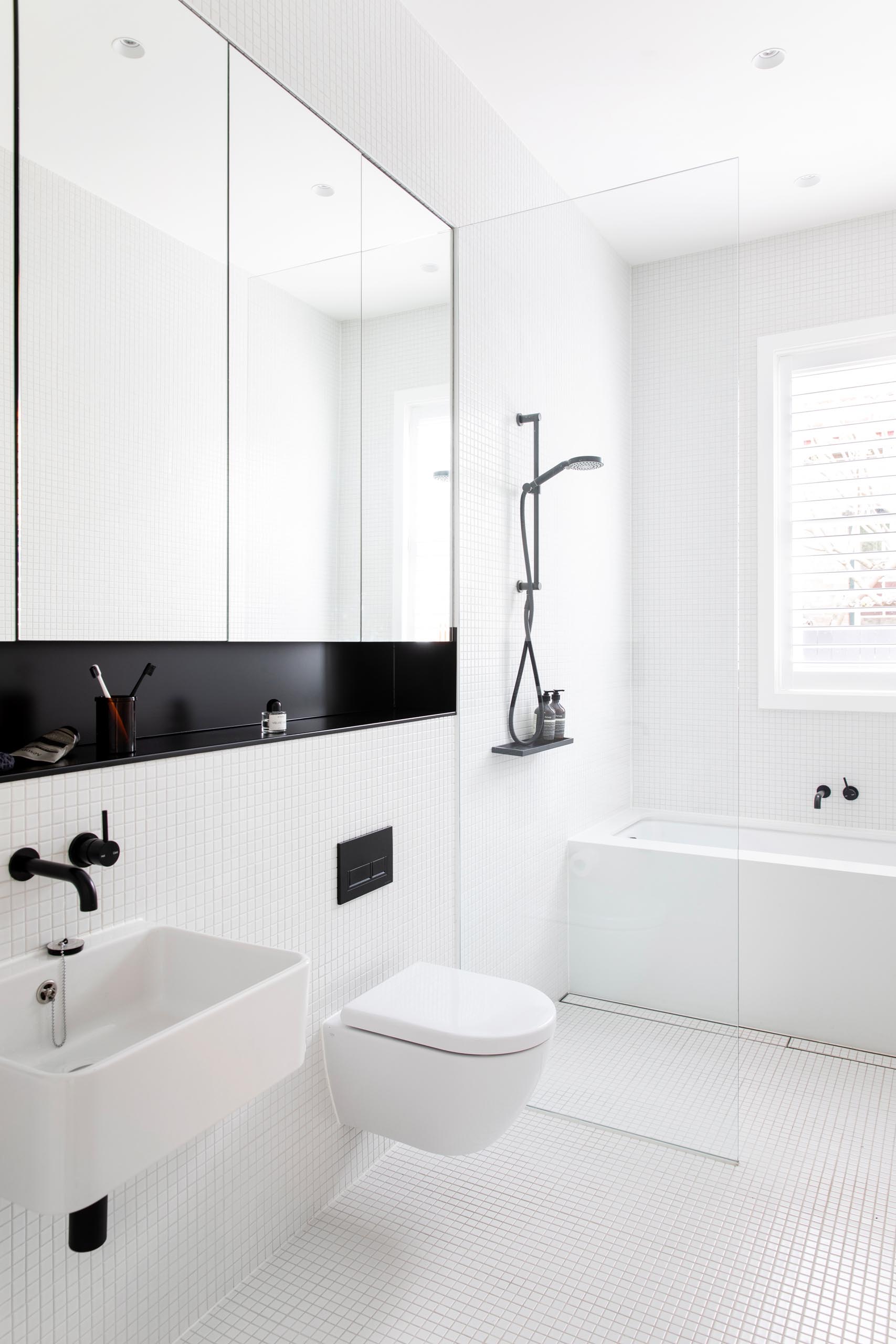 This modern bathroom has a simple white design with black accents. There's small white square tiles covering the walls and floor, a built-in bathtub, and a mirror with a recessed black shelf below. In addition to the window above the bath, there's also a skylight to add natural light to the space.