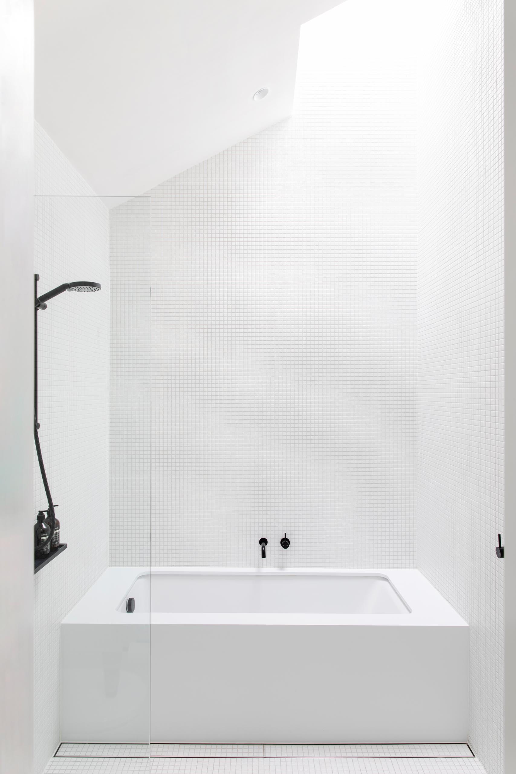 A white bathroom with black hardware accents features small square tiles, a built-in bathtub, and a skylight.