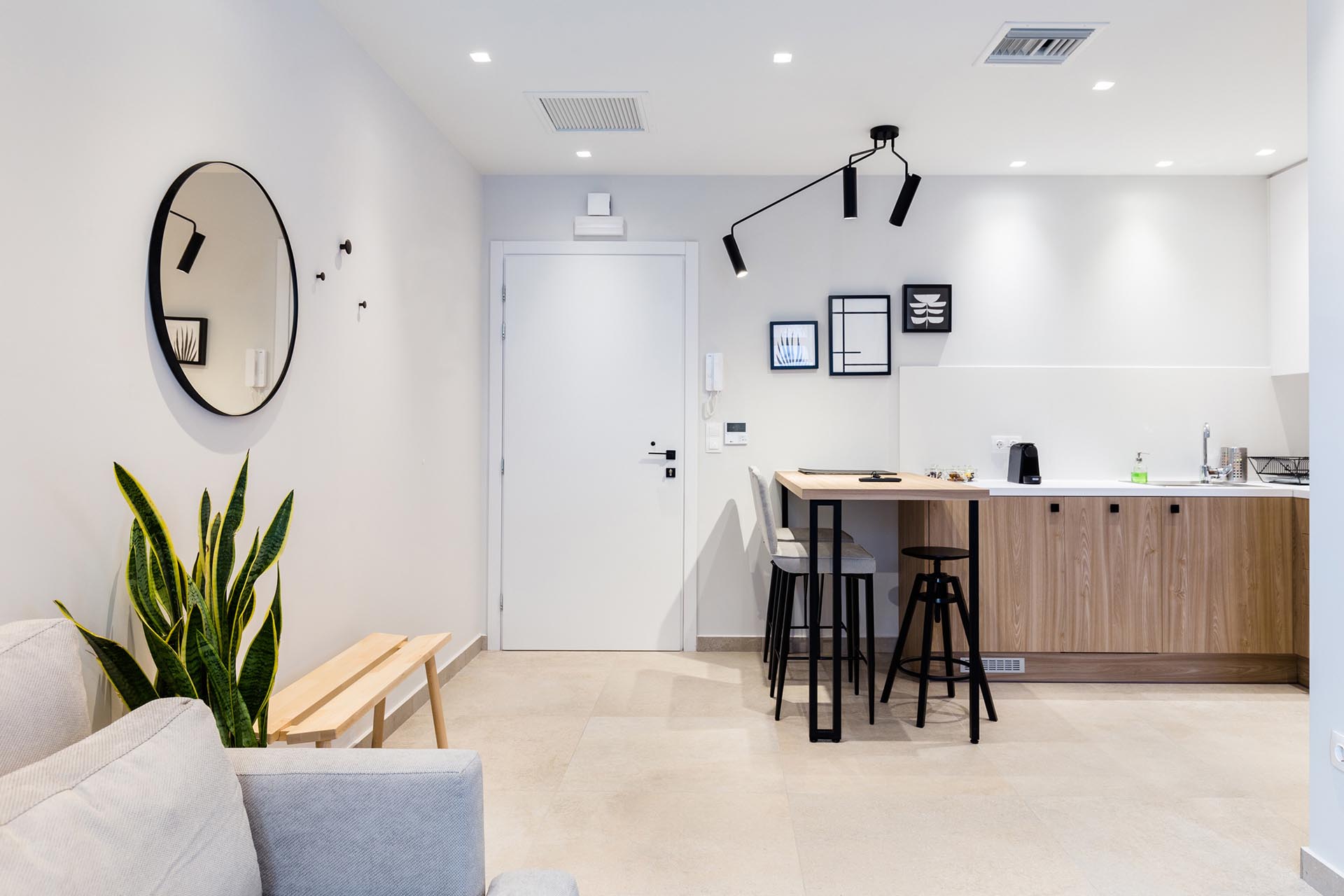 This small apartment has an open interior with a kitchen that features minimalist white upper cabinets and wood lower cabinets, as well as a bar height table.