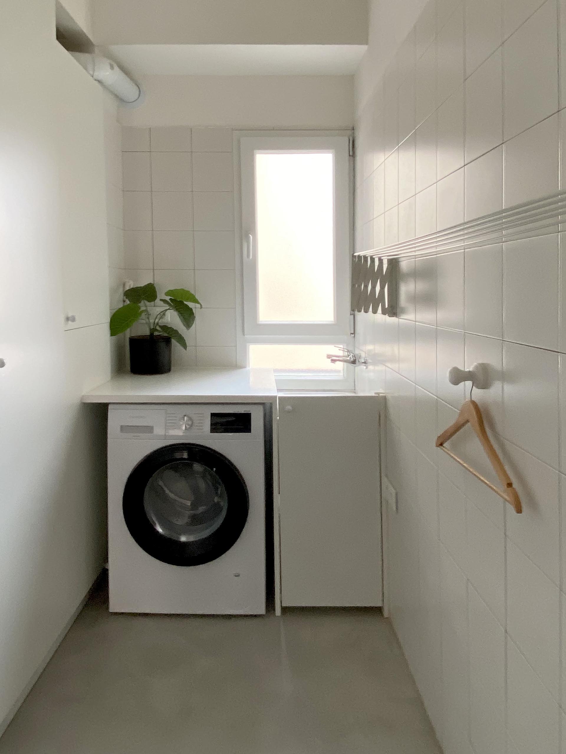 A small laundry with square white tiles and a sink below the window.