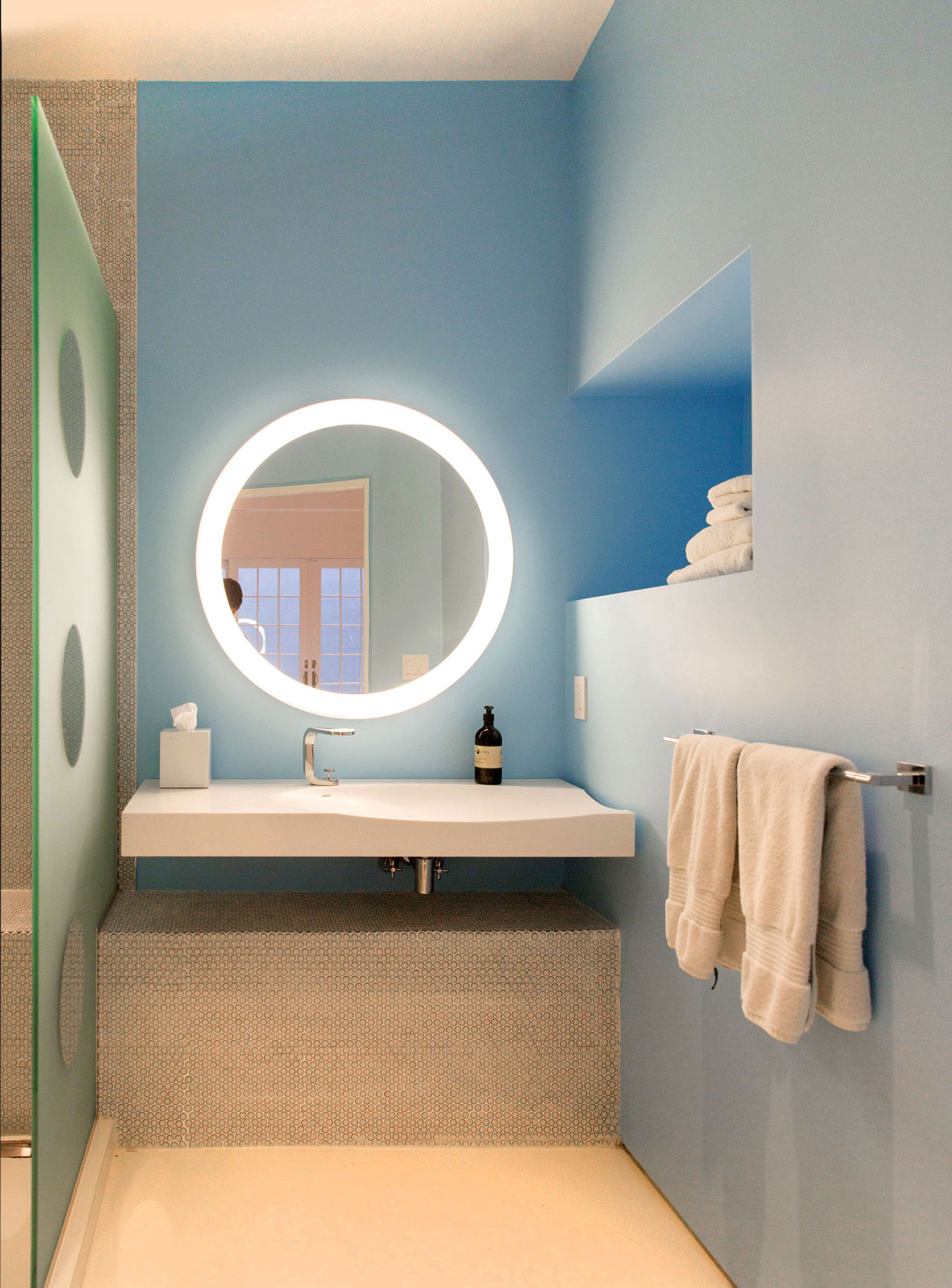 A basement bathroom that features design elements like small penny tiles, a translucent glass shower screen with clear circular windows, and a round vanity mirror with integrated lighting.