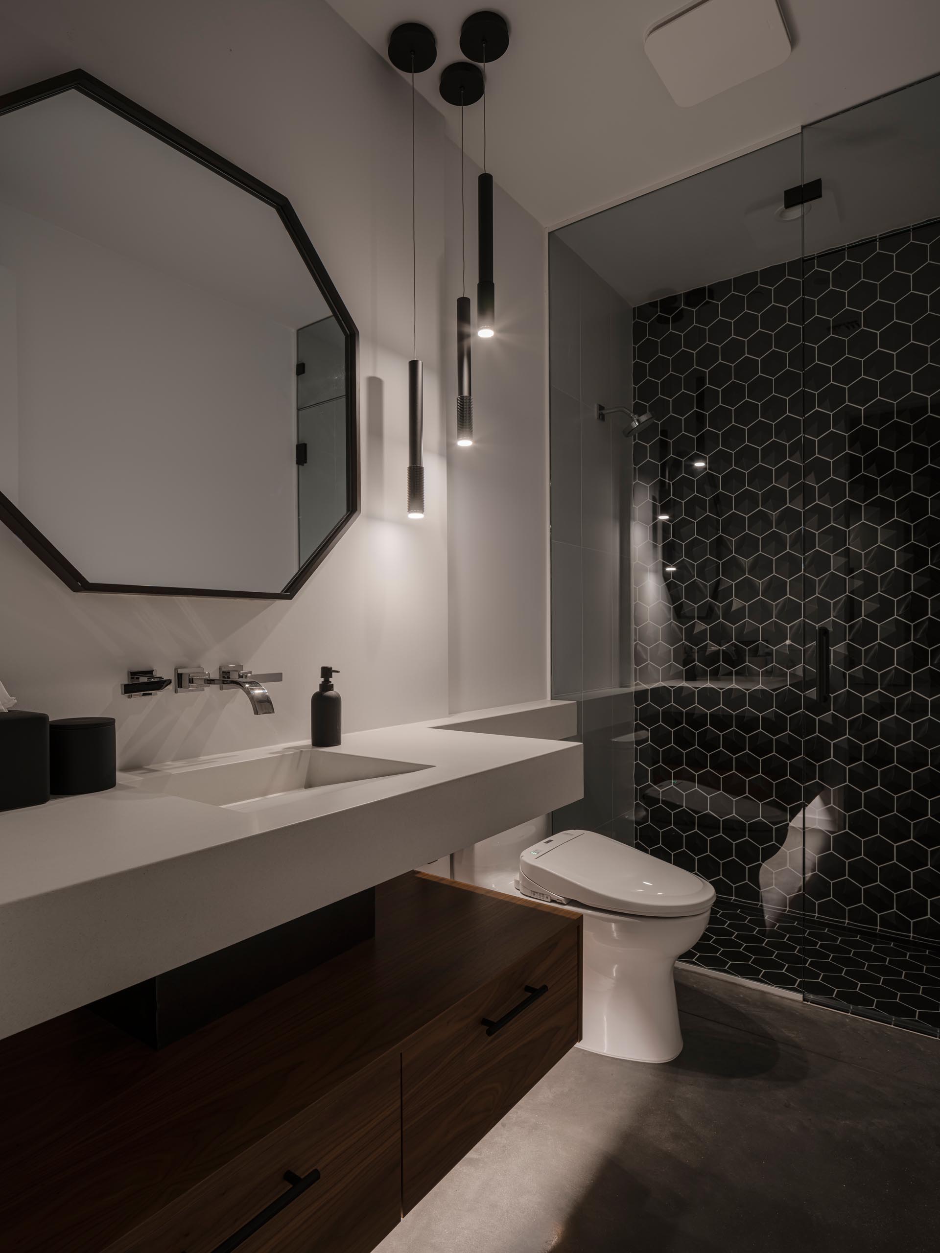 In this modern guest bathroom there's a floating vanity with wood cabinet underneath, and a large black framed octagonal mirror that complements the black hexagonal tiles in the shower.