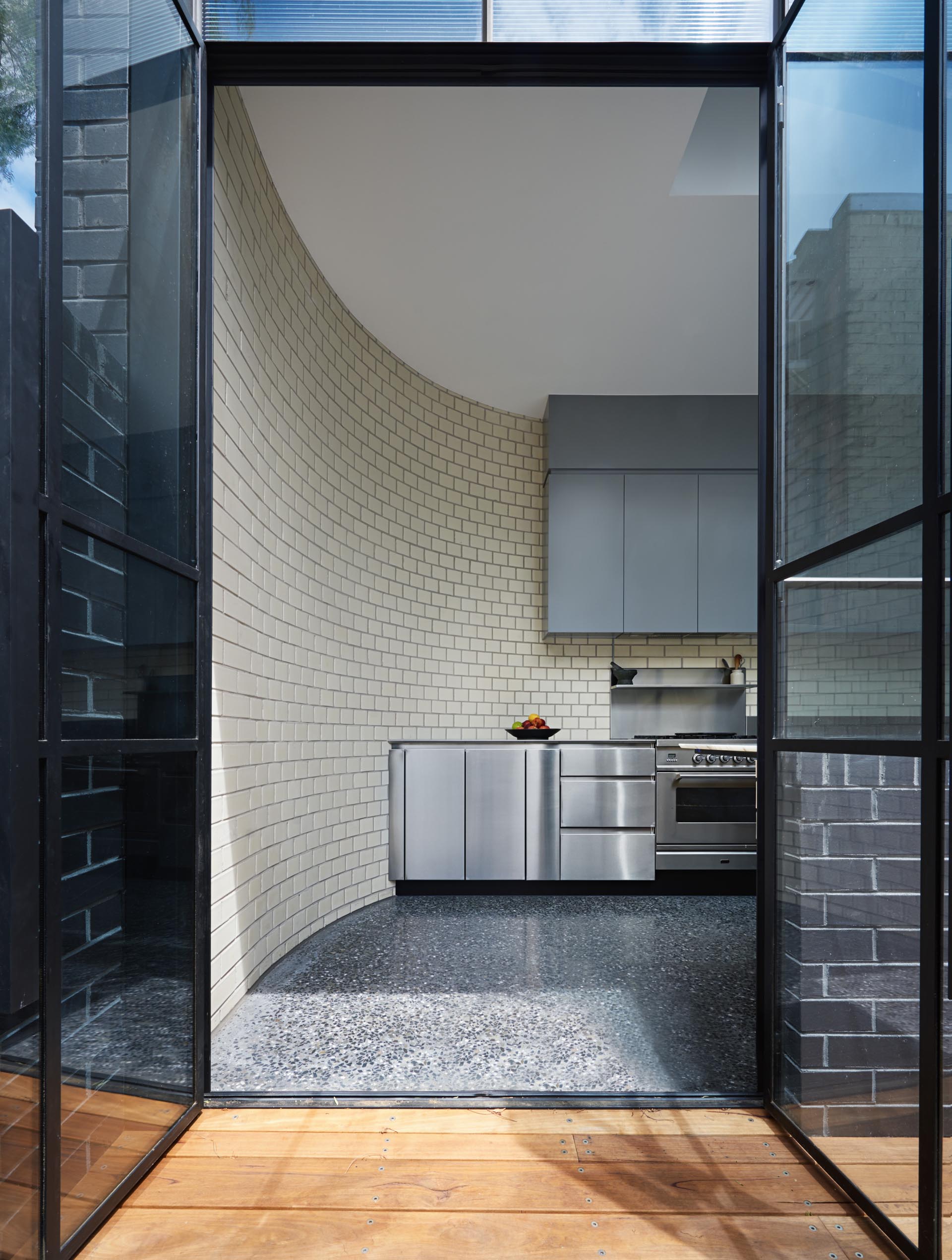 This modern kitchen is designed with a curved white brick wall, stainless steel lower cabinets, and minimalist upper cabinets with a matte finish. Opposite this wall of cabinetry, is a long timber countertop with built-in marble section for kneading dough.