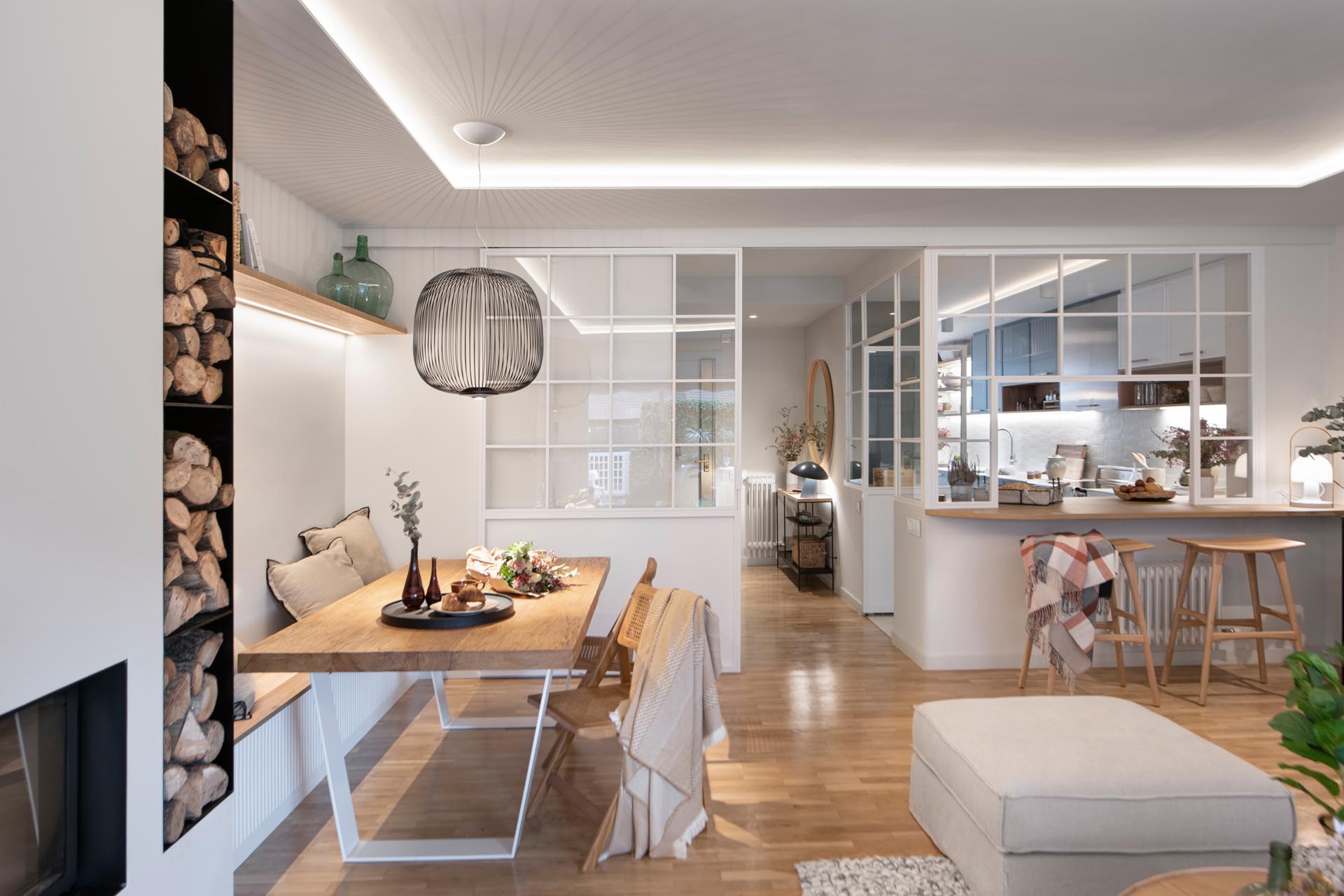 This open plan dining area includes a long bench against the wall, a wood table with wood chairs, a pendant light that gives of lined shadows, and a wood shelf with hidden lighting underneath.