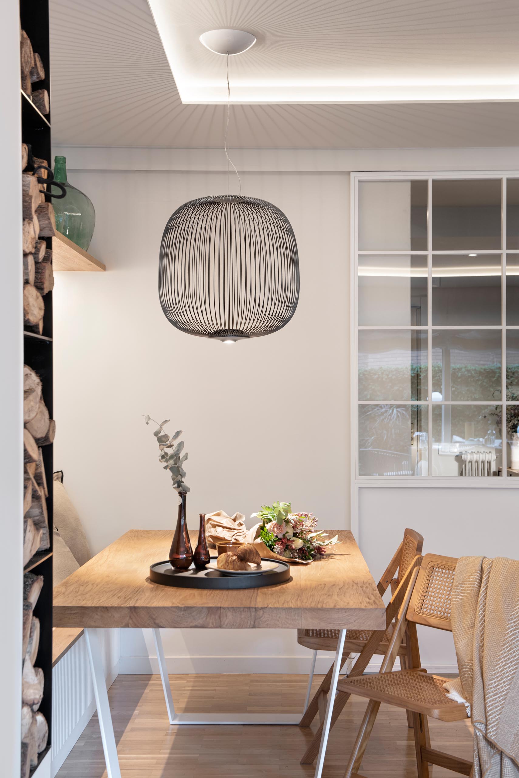 This open plan dining area includes a long bench against the wall, a wood table with wood chairs, a pendant light that gives of lined shadows, and a wood shelf with hidden lighting underneath.