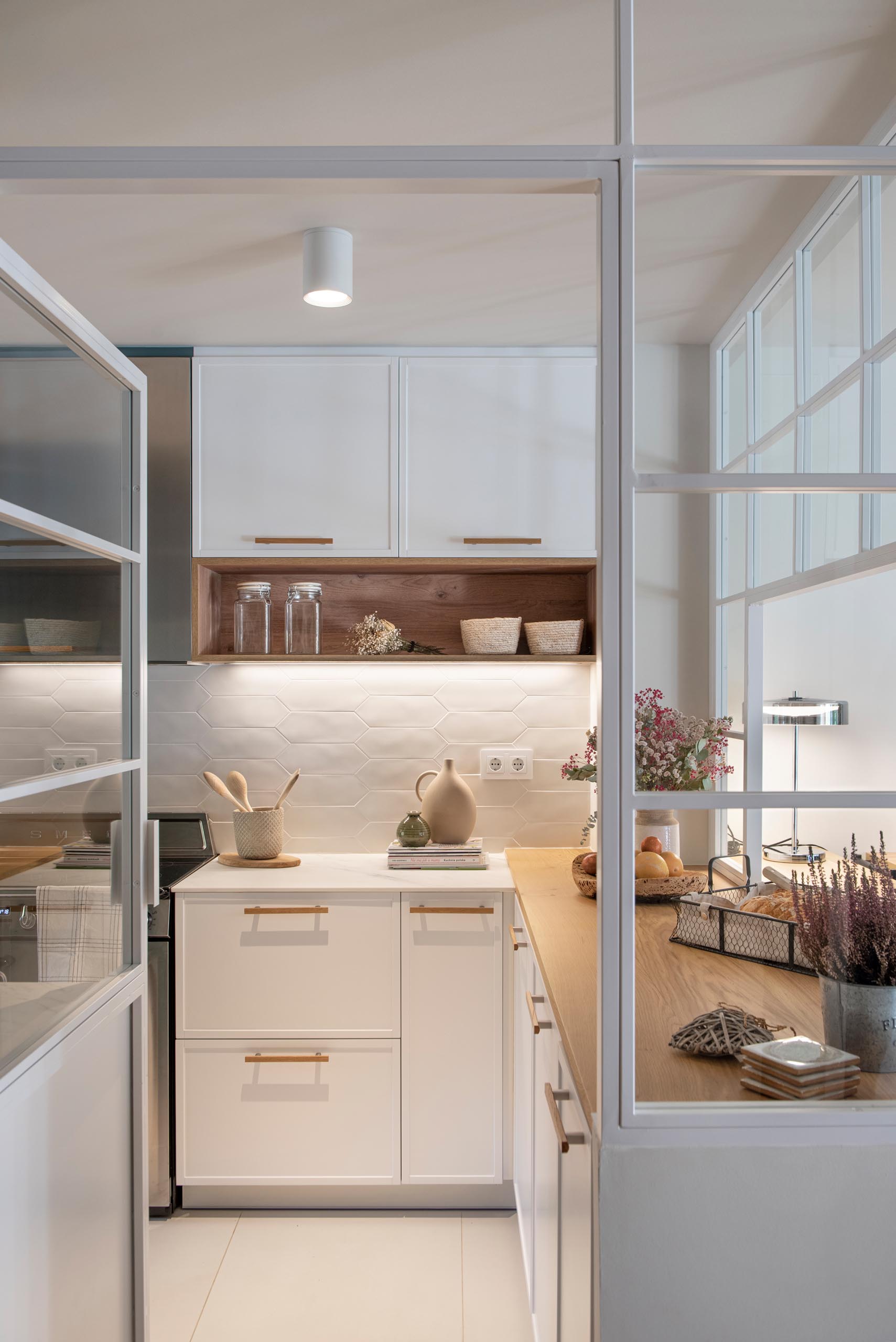 A modern kitchen enclosed within white-framed windows, includes white cabinets that have been paired with a matte green cabinets, while white tiles have been used for the backsplash. An eat-in breakfast nook is tucked into the corner and includes a built-in bench, and a mirrored wall, which makes the space feel larger.