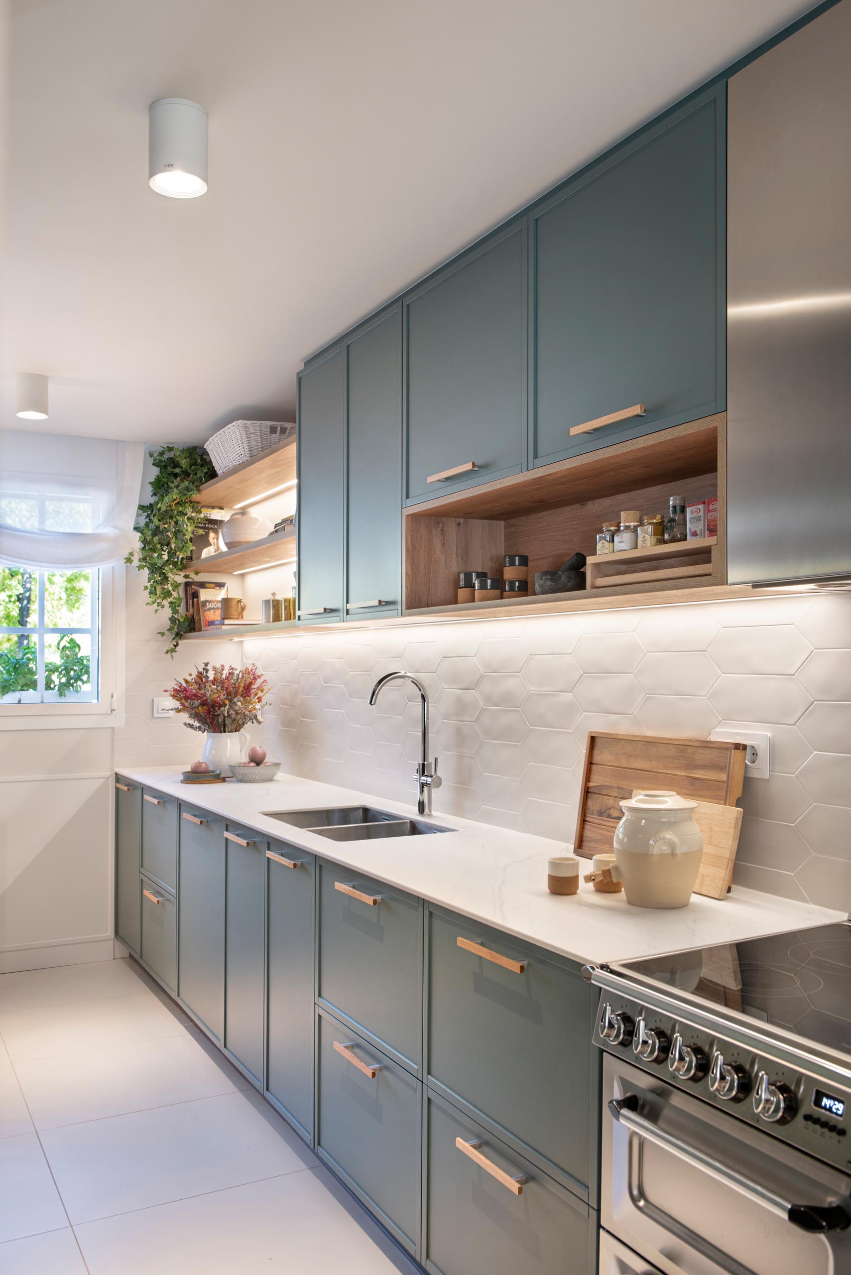 A modern kitchen enclosed within white-framed windows, includes white cabinets that have been paired with a matte green cabinets, while white tiles have been used for the backsplash. An eat-in breakfast nook is tucked into the corner and includes a built-in bench, and a mirrored wall, which makes the space feel larger.