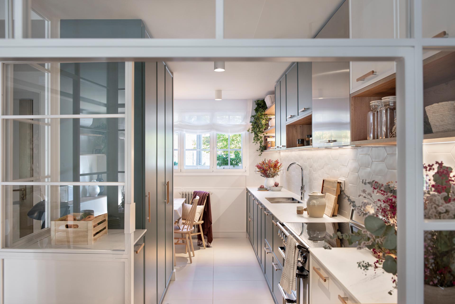 A modern kitchen enclosed within white-framed windows, includes white cabinets that have been paired with a matte green cabinets, while white tiles have been used for the backsplash. An eat-in breakfast nook is tucked into the corner and includes a built-in bench, and a mirrored wall, which makes the space feel larger.