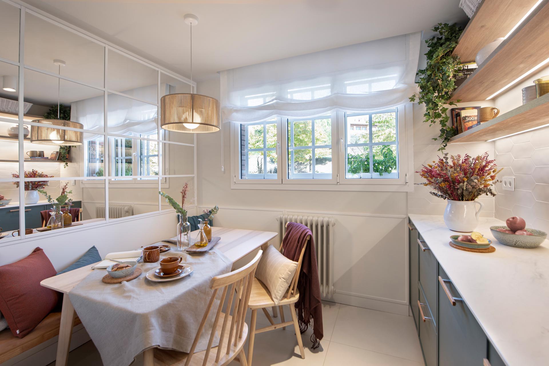 A modern kitchen enclosed within white-framed windows, includes white cabinets that have been paired with a matte green cabinets, while white tiles have been used for the backsplash. An eat-in breakfast nook is tucked into the corner and includes a built-in bench, and a mirrored wall, which makes the space feel larger.