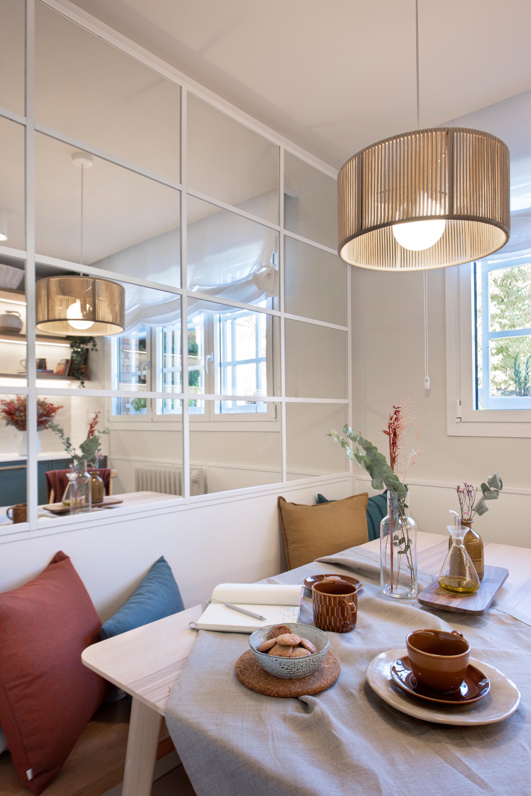 A modern kitchen enclosed within white-framed windows, includes white cabinets that have been paired with a matte green cabinets, while white tiles have been used for the backsplash. An eat-in breakfast nook is tucked into the corner and includes a built-in bench, and a mirrored wall, which makes the space feel larger.