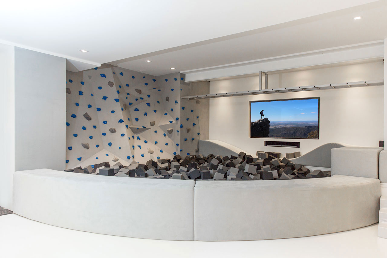 This modern kid's playroom in a basement includes a foam pit area that's defined by a upholstered low wall with a curved shape. This low wall encloses the foam pieces, which act as a soft area to land, should a child fall or jump from the rock climbing wall or monkey bar.