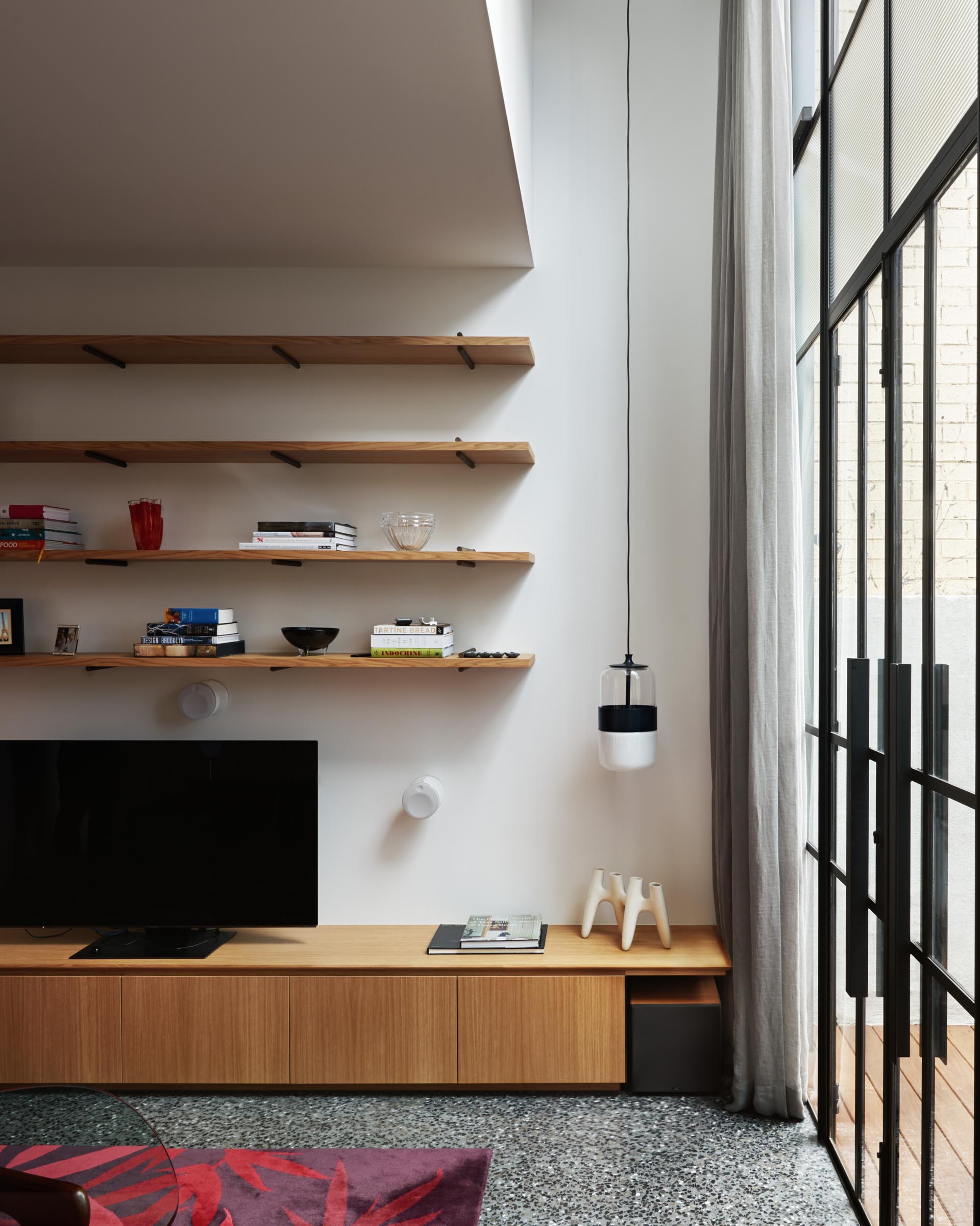 The new living room includes a wood storage unit that acts as a TV stand, as well as multiple long wood shelves that make use of the wall. The floor is a polished concrete with under-floor heating for the cooler temperatures.