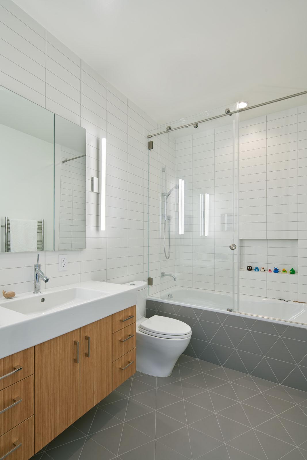 In this modern bathroom, simple white rectangular tiles cover the walls, while gray triangle tiles cover the floor and wrap onto the bathtub.