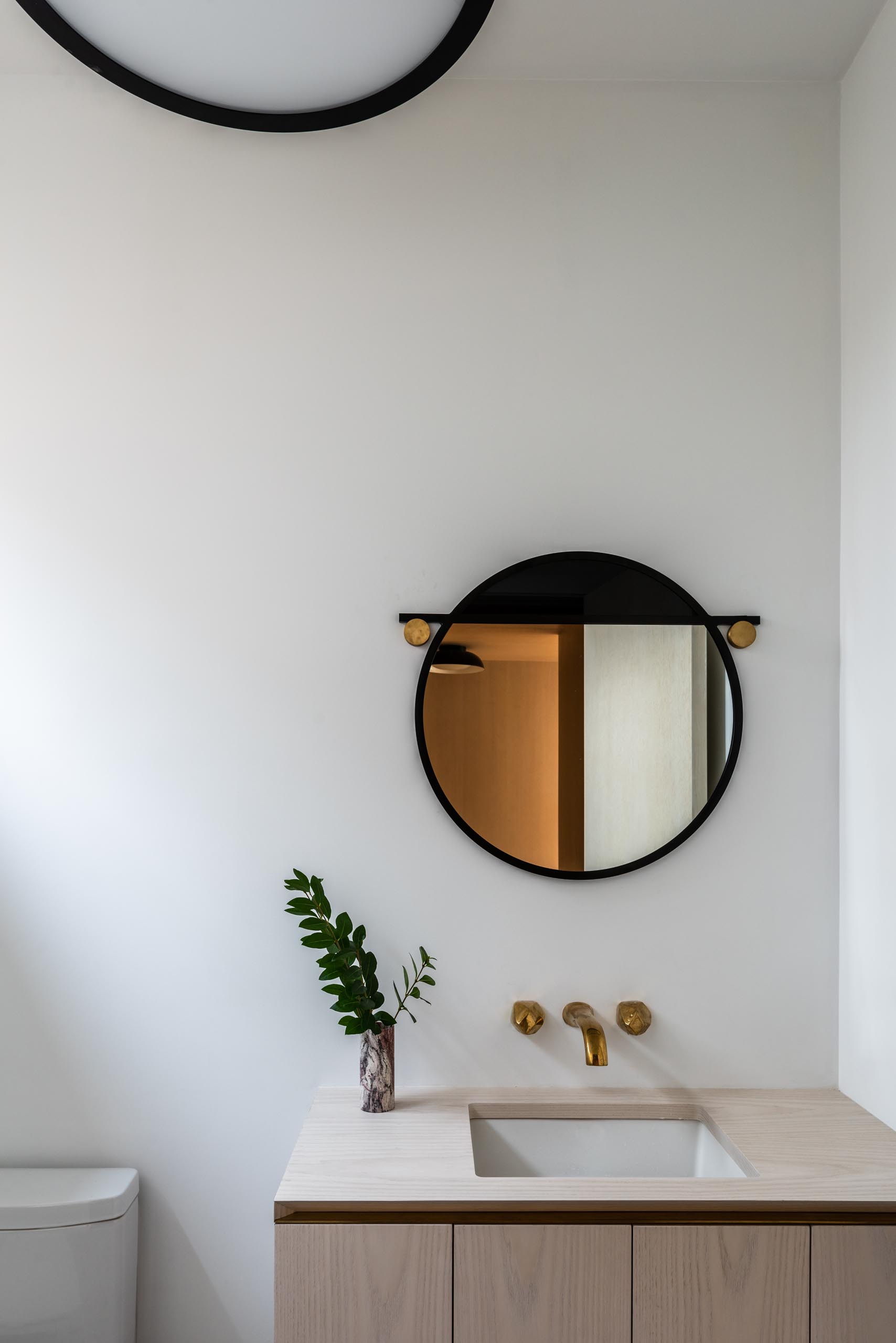 In this modern bathroom, the focus is the round mirror with a thick black frame that hangs above a light wood vanity with an undermount sink.