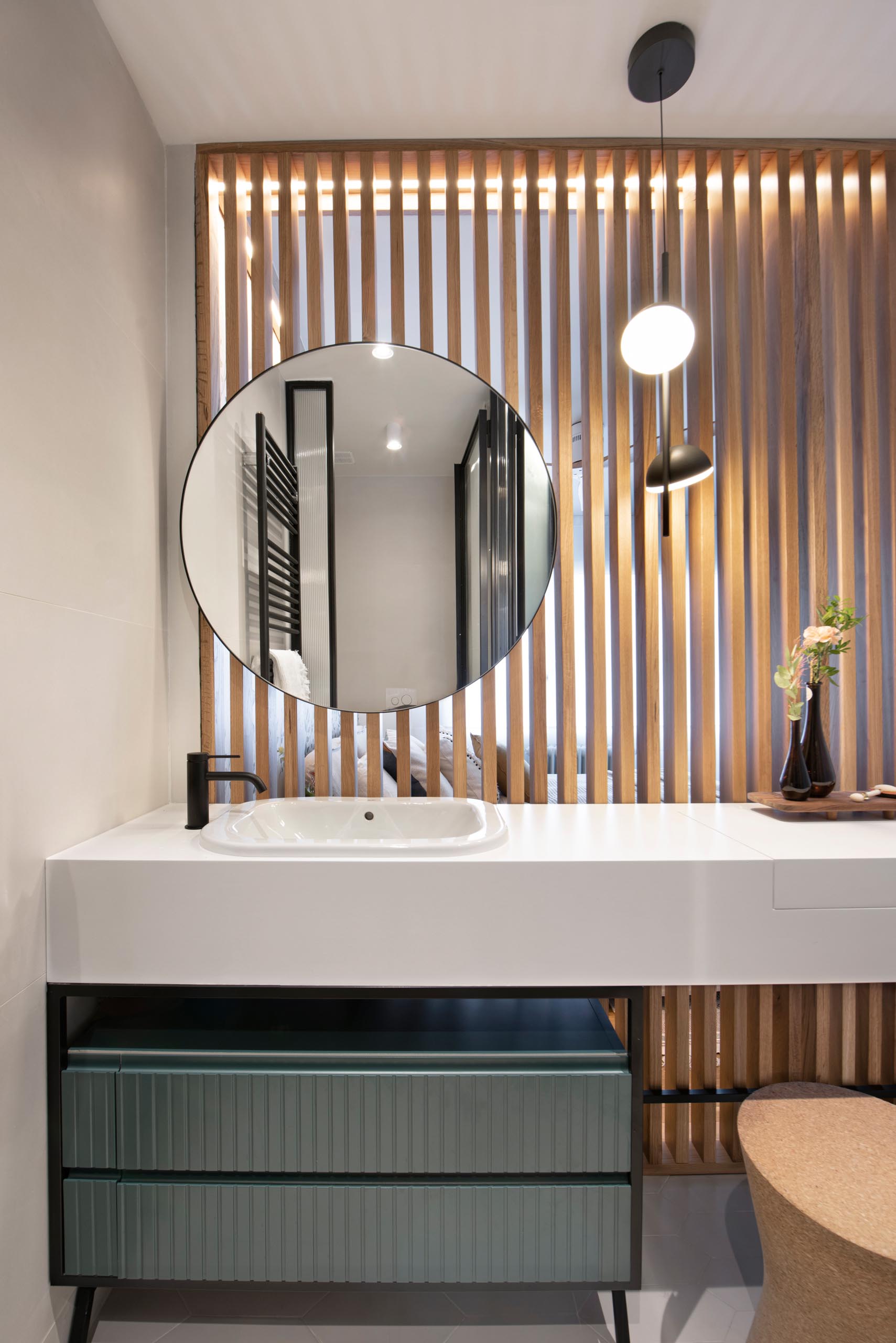 This en-suite bathroom is separated from the bedroom by a wood slat wall, while black framed glass walls hide the shower and toilet, and the vanity has a thick white countertop that hides a makeup area.