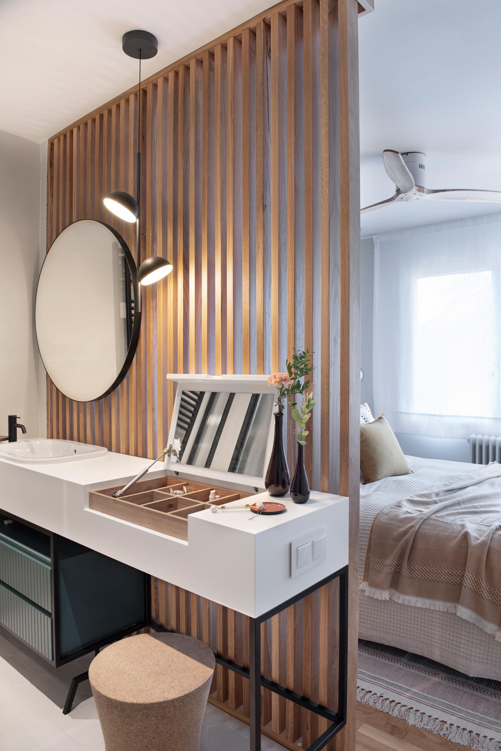 This en-suite bathroom is separated from the bedroom by a wood slat wall, while black framed glass walls hide the shower and toilet, and the vanity has a thick white countertop that hides a makeup area.