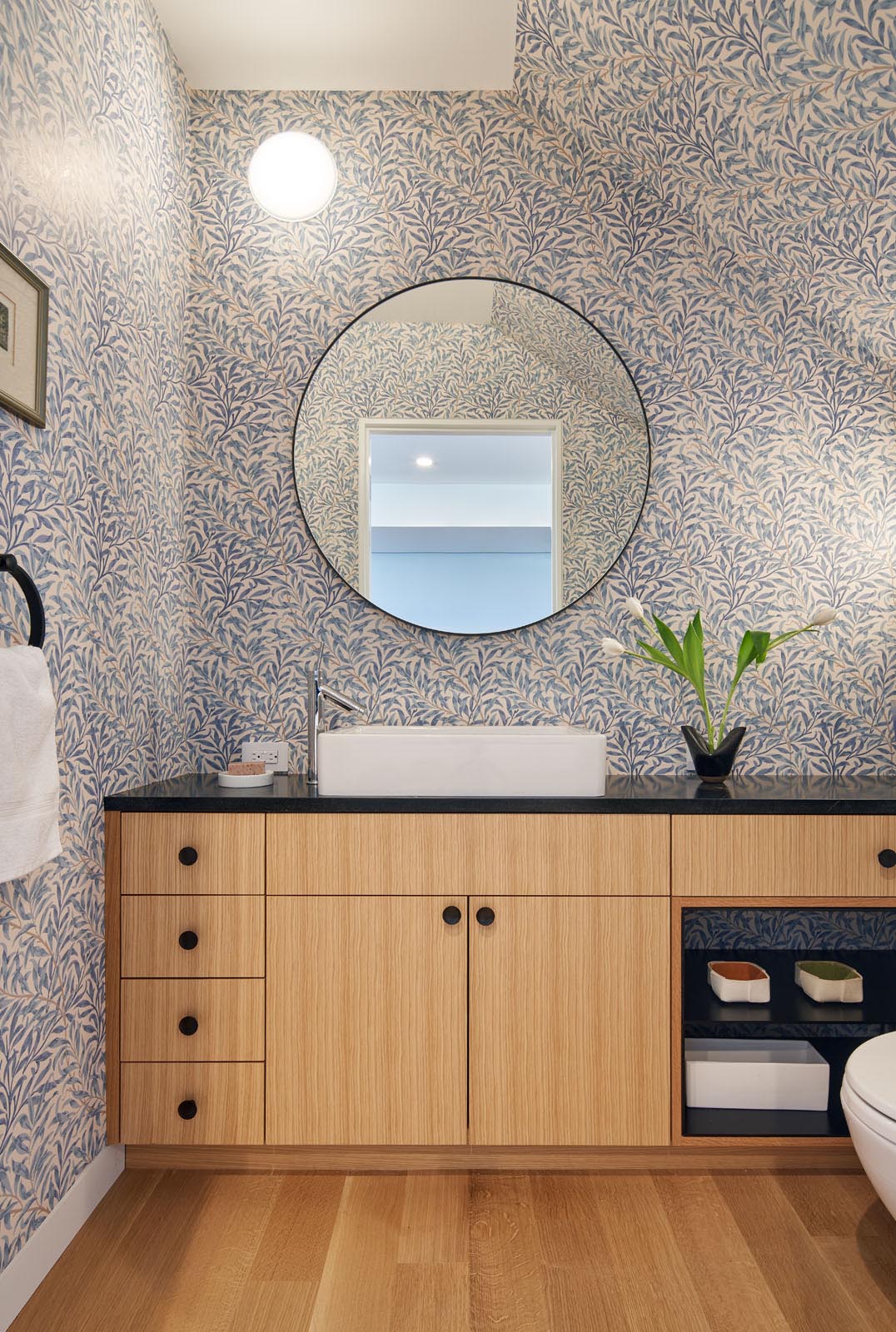 In this modern bathroom, the walls are covered in a wallpaper with a leaf design, while a round mirror hangs above a wood vanity with a black countertop.