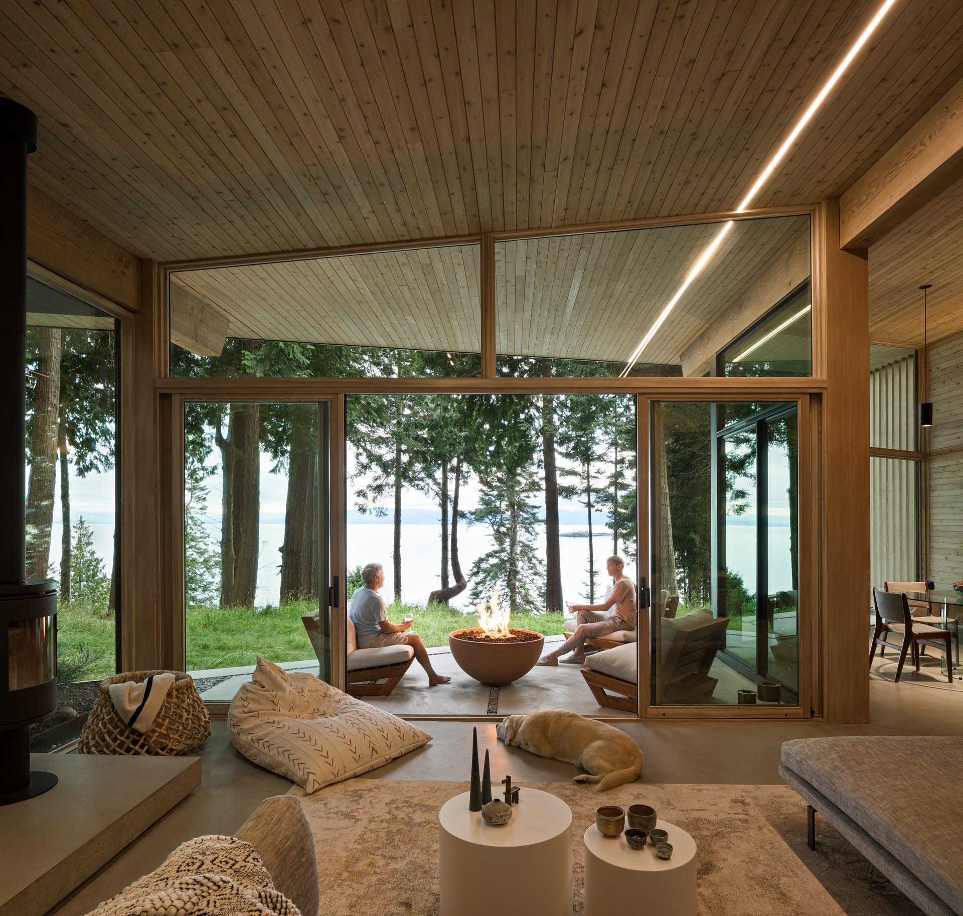 A wall of glass with sliding doors connects the living room to a covered patio that's furnished with chairs and a fire bowl, and has water views.