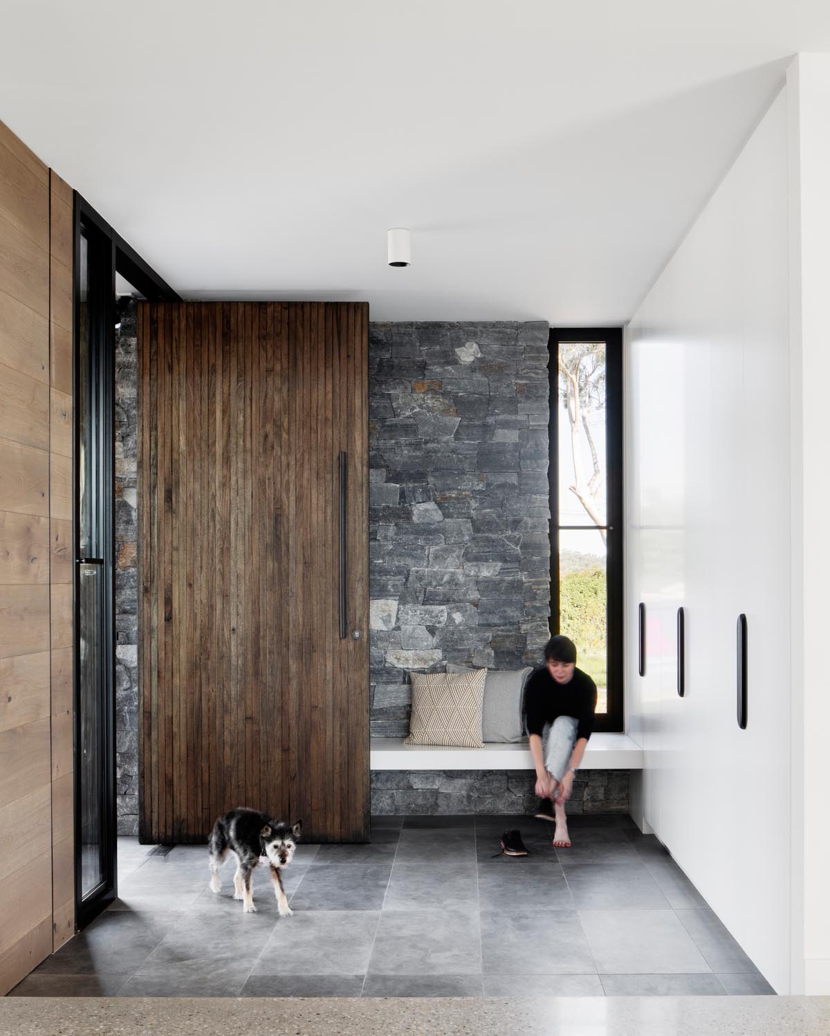 A large front door with dark vertical wood welcomes visitors to the home, with the stone wall continuing through the interior. A wall of floor-to-ceiling white cabinets provides plenty of storage, while a bench creates a place to take shoes off.