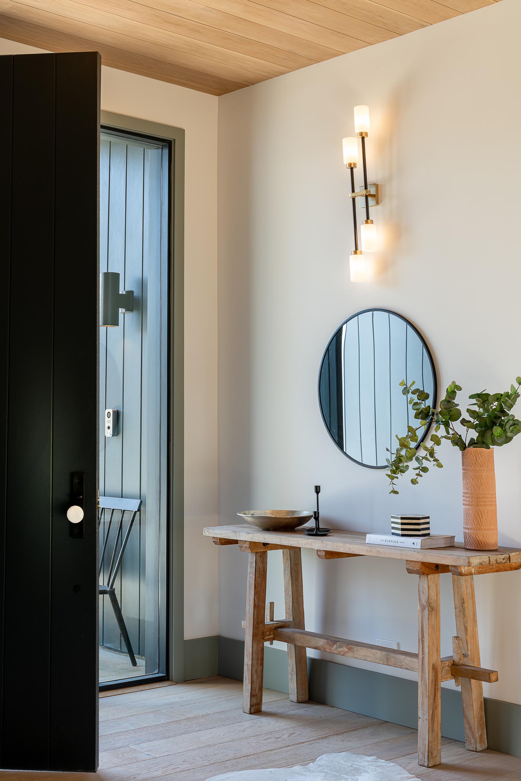 This entryway has been kept simple with a rustic wood console table, a round mirror, a wall sconce, and white oak wide plank flooring.