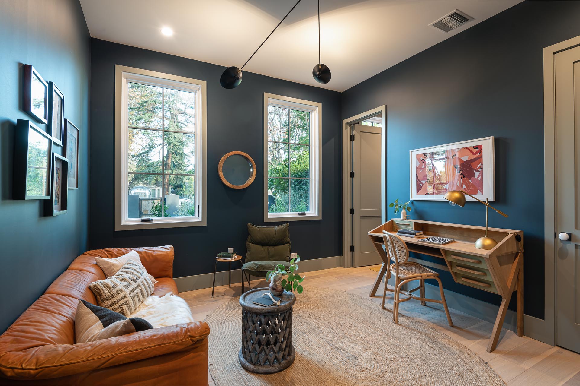 A modern home office with white framed windows, dark walls, a leather sofa, and a wood desk.