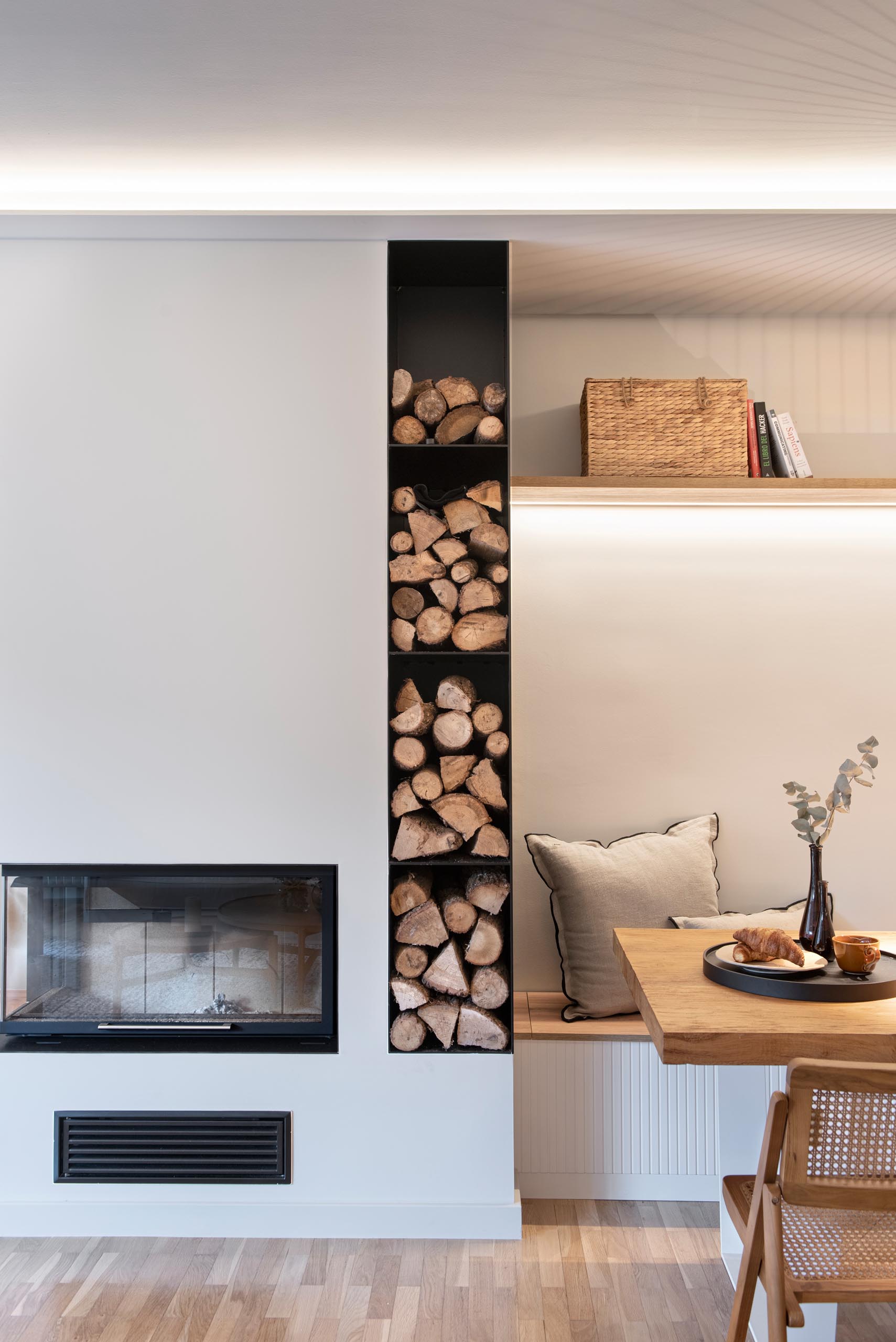 This modern open plan living room has shelving that lines the wall, while the couch and armchair are focused on the fireplace and built-in firewood storage.