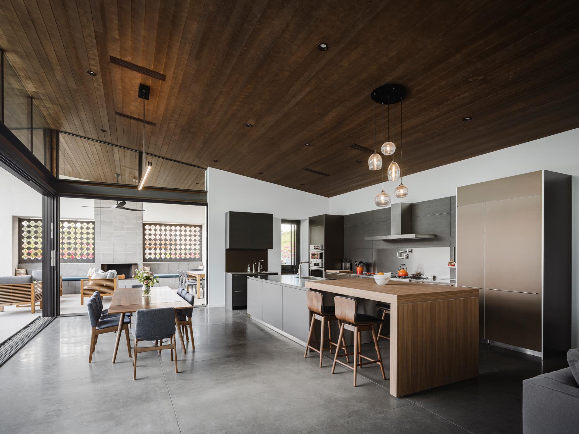 The open plan dining room and kitchen of this modern home includes concrete flooring, a wood dining table, and a large kitchen with an expansive island with a raised section that acts as a bar.
