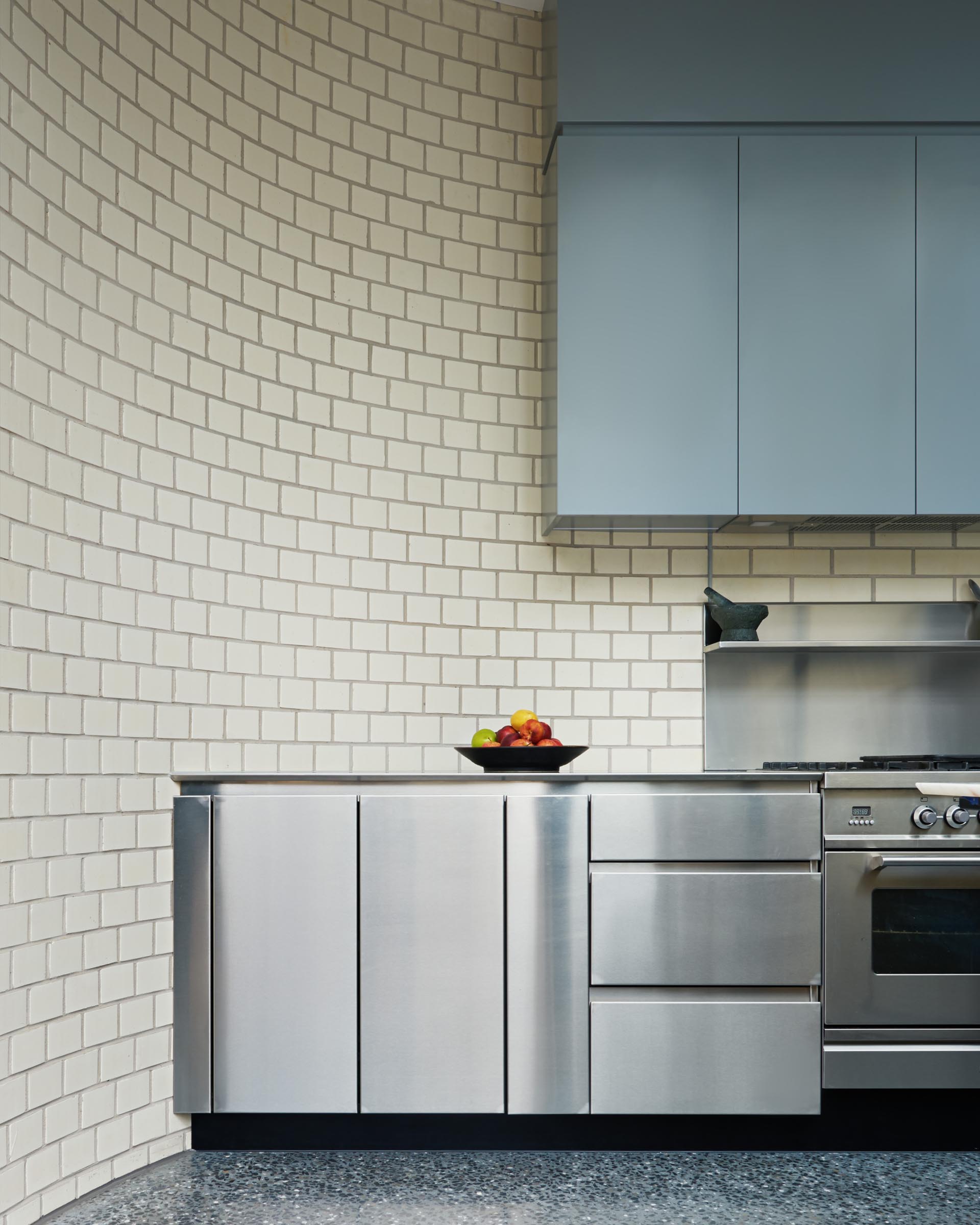 This modern kitchen is designed with a curved white brick wall, stainless steel lower cabinets, and minimalist upper cabinets with a matte finish. Opposite this wall of cabinetry, is a long timber countertop with built-in marble section for kneading dough.