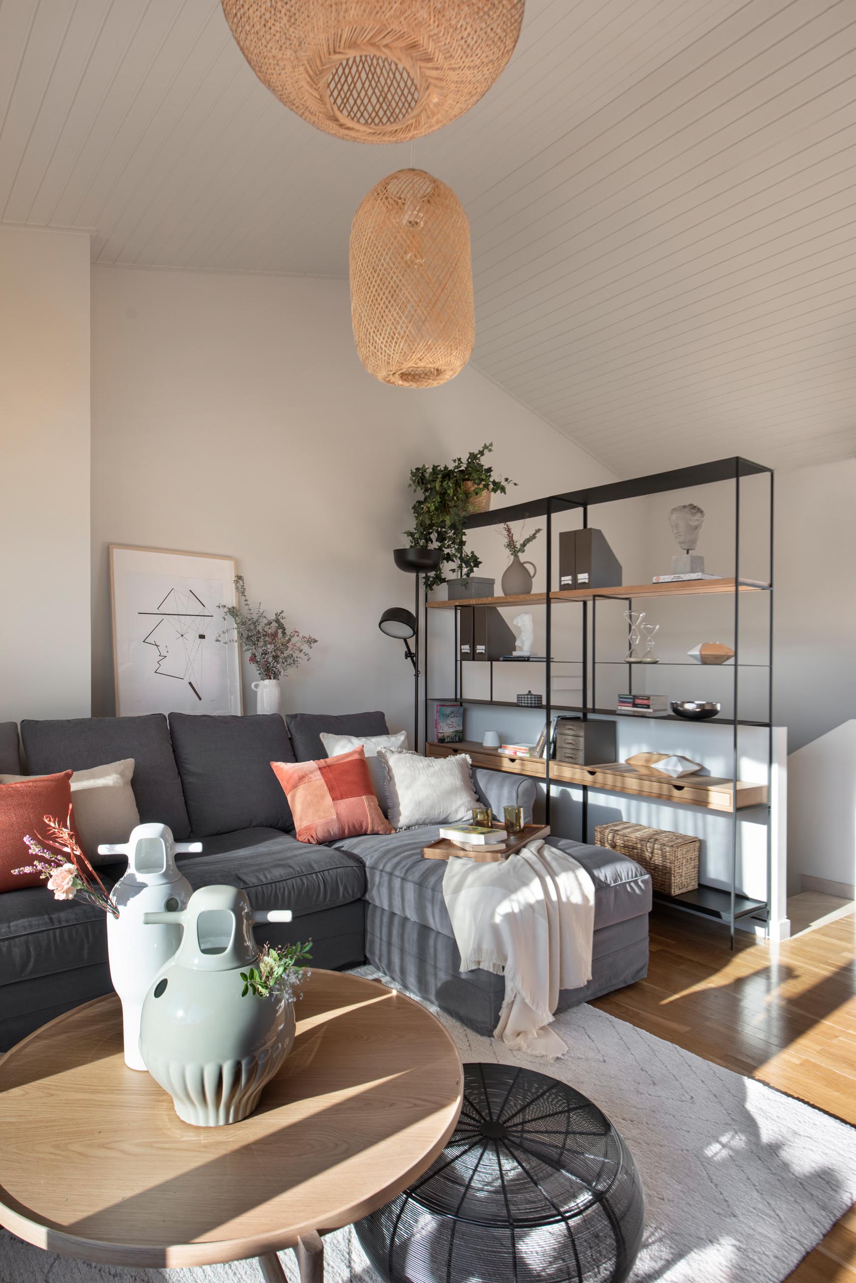 On the top level of this modern home is a secondary living room with a pitched ceiling. The living room is furnished with a couch, shelving unit, rattan pendant lights, a desk with TV above it, and a reading area with built-in shelves.