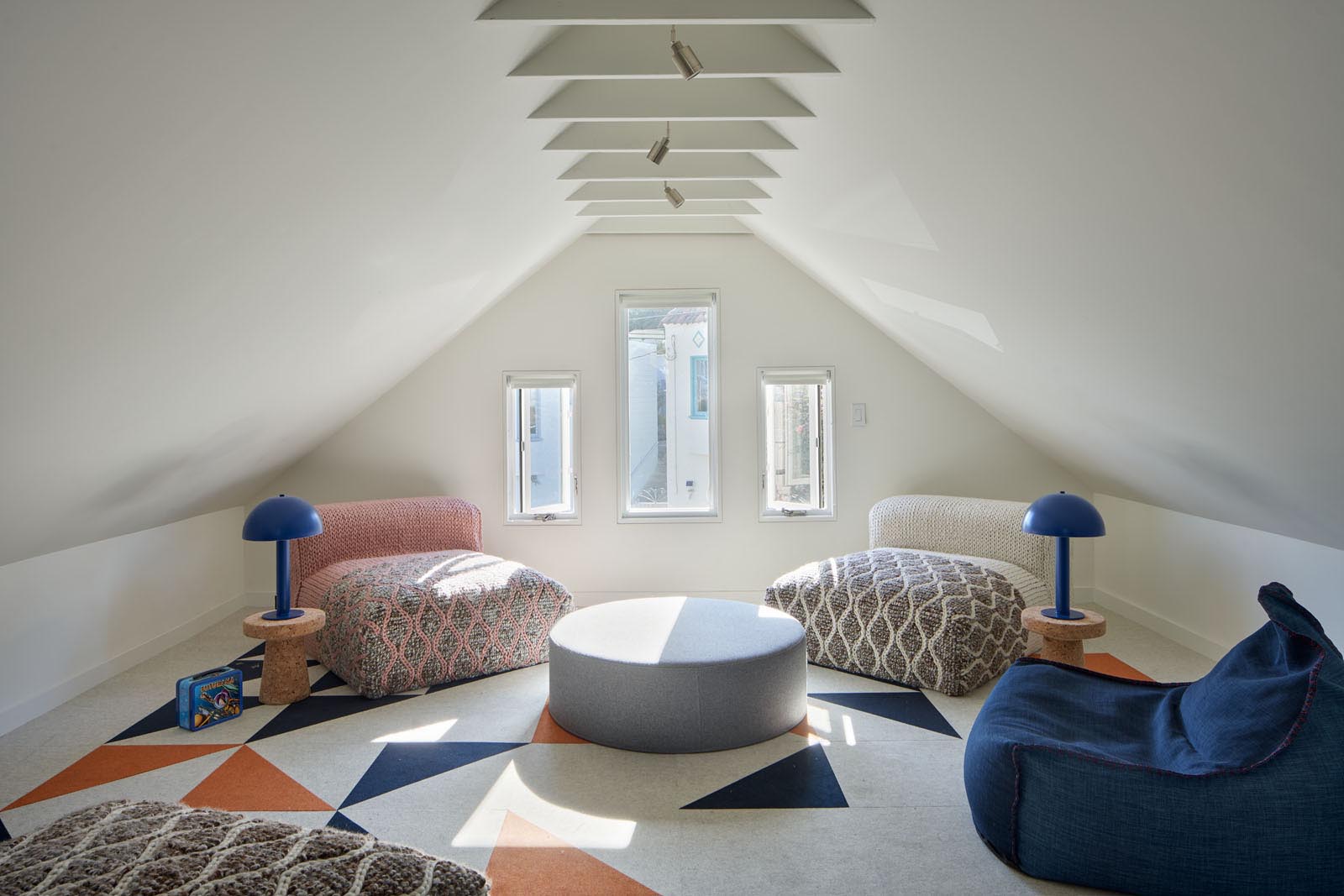 A modern attic that's been furnished with comfortable low chairs, a pair of lights, a rug, and an upholstered round ottoman.