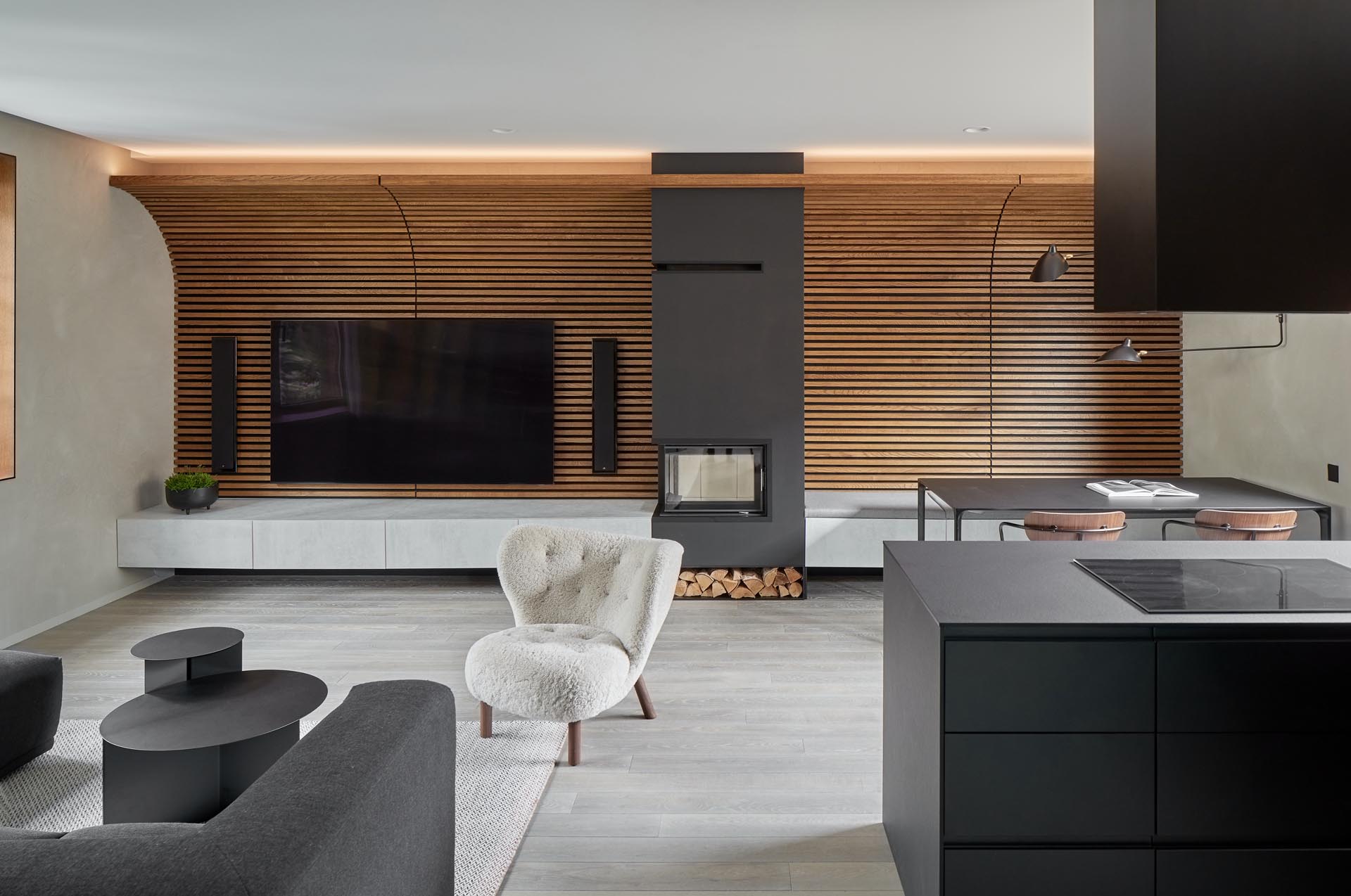 A wood slat accent wall is curved towards the ceiling, and becomes the backdrop for the television and wall-mounted speakers. A fireplace with a black surround that incorporates firewood storage, separates the living room side from the dining area.