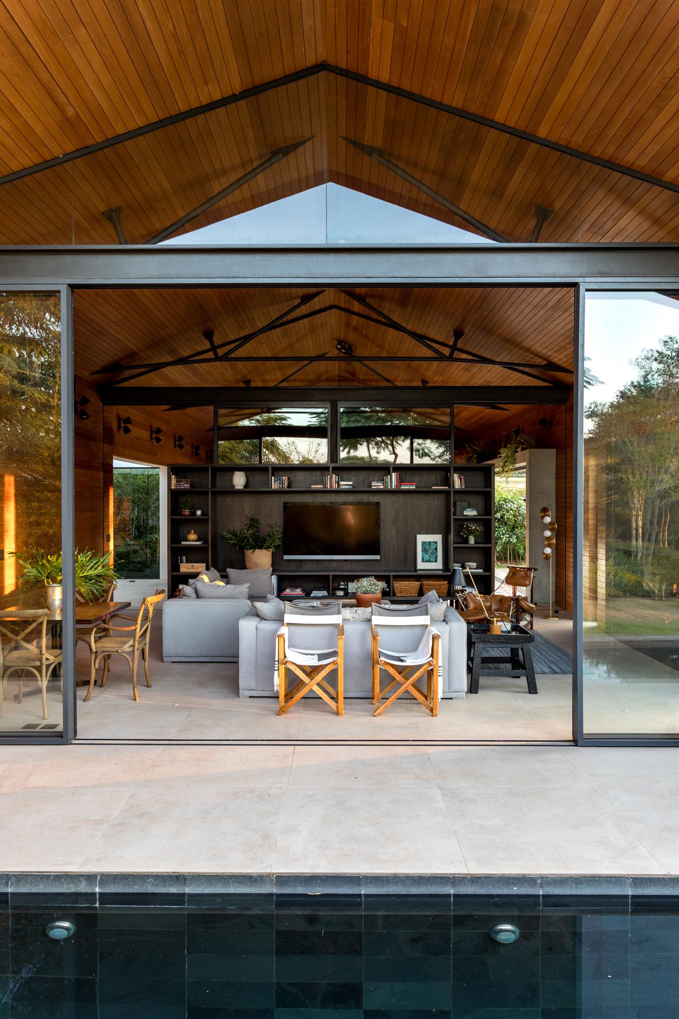 This modern living room is open to the swimming pool, and is furnished with a pair of couches, armchairs, and a fireplace. The room divider is a mirror image of the other side, with shelving and a mounted TV.