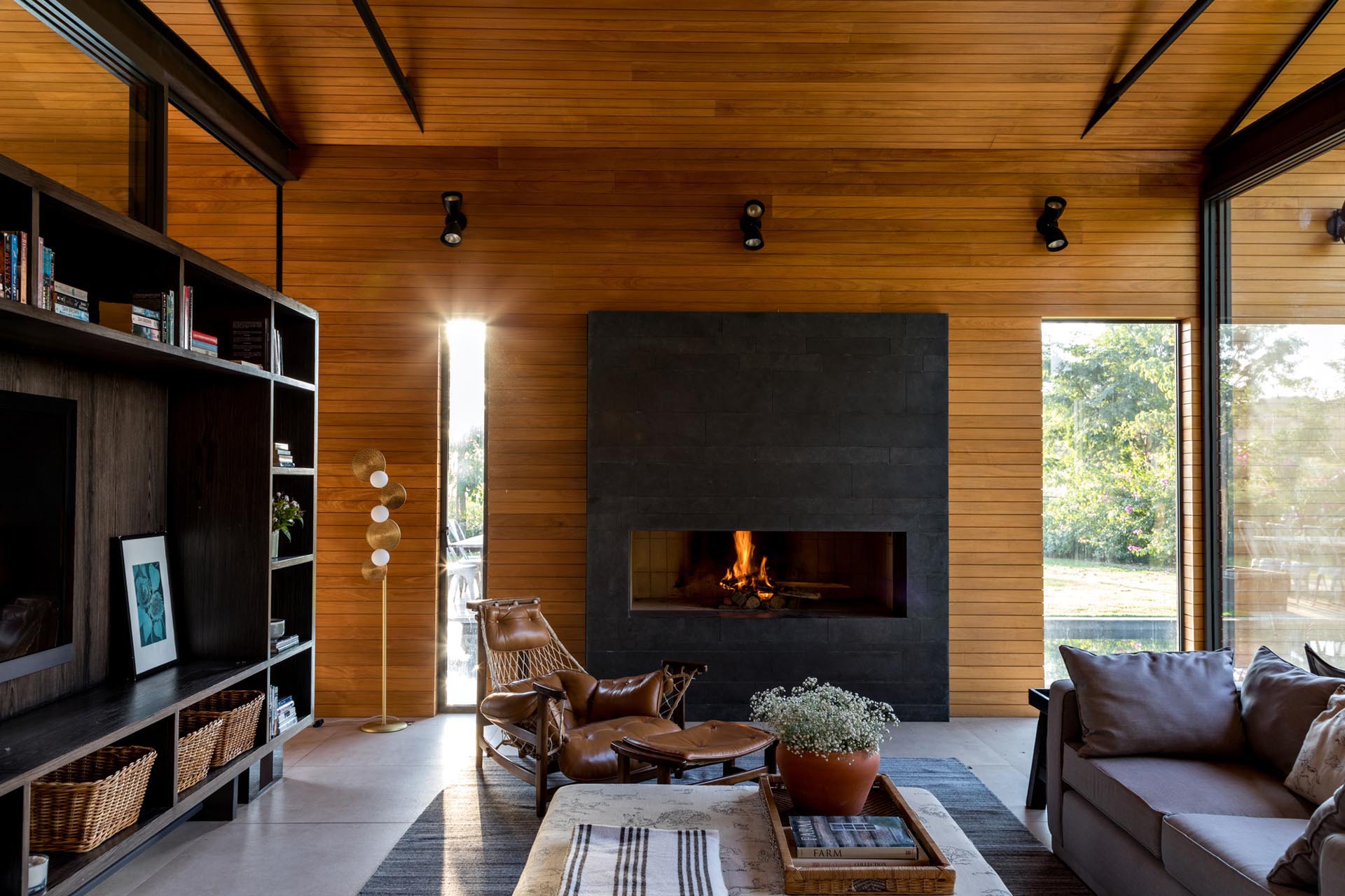 This modern living room is open to the swimming pool, and is furnished with a pair of couches, armchairs, and a fireplace. The room divider is a mirror image of the other side, with shelving and a mounted TV.