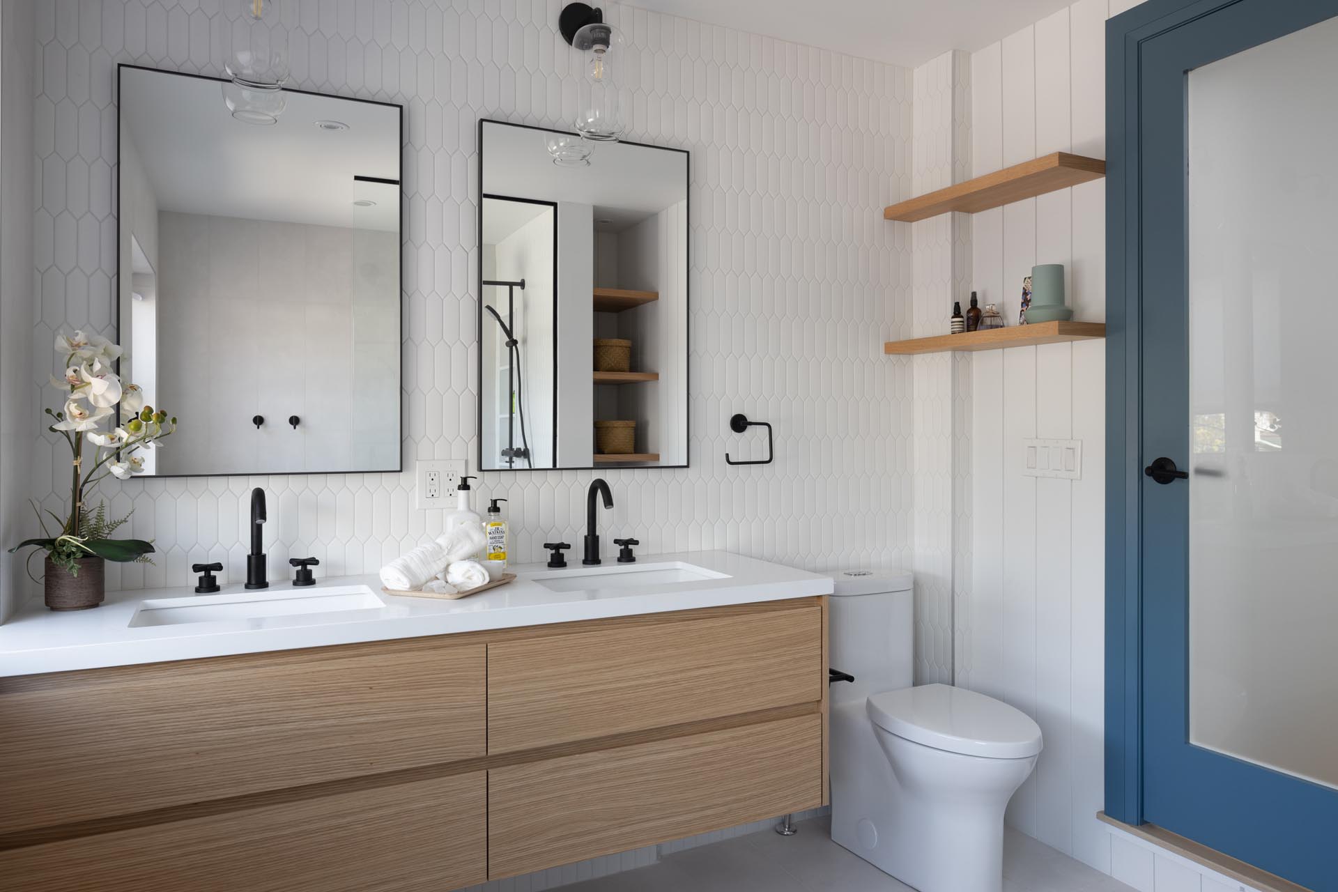 This modern bathroom includes a picket-style tile installation on the wall, a wood vanity with double sinks and black framed mirrors.