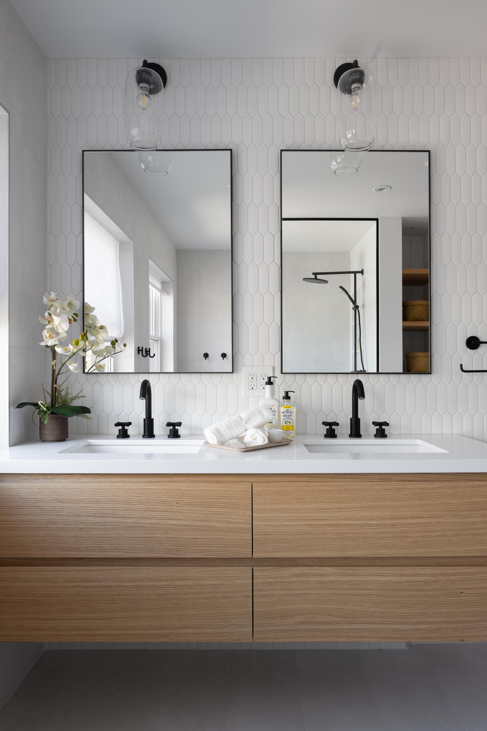 This modern bathroom includes a picket-style tile installation on the wall, a wood vanity with double sinks and black framed mirrors.