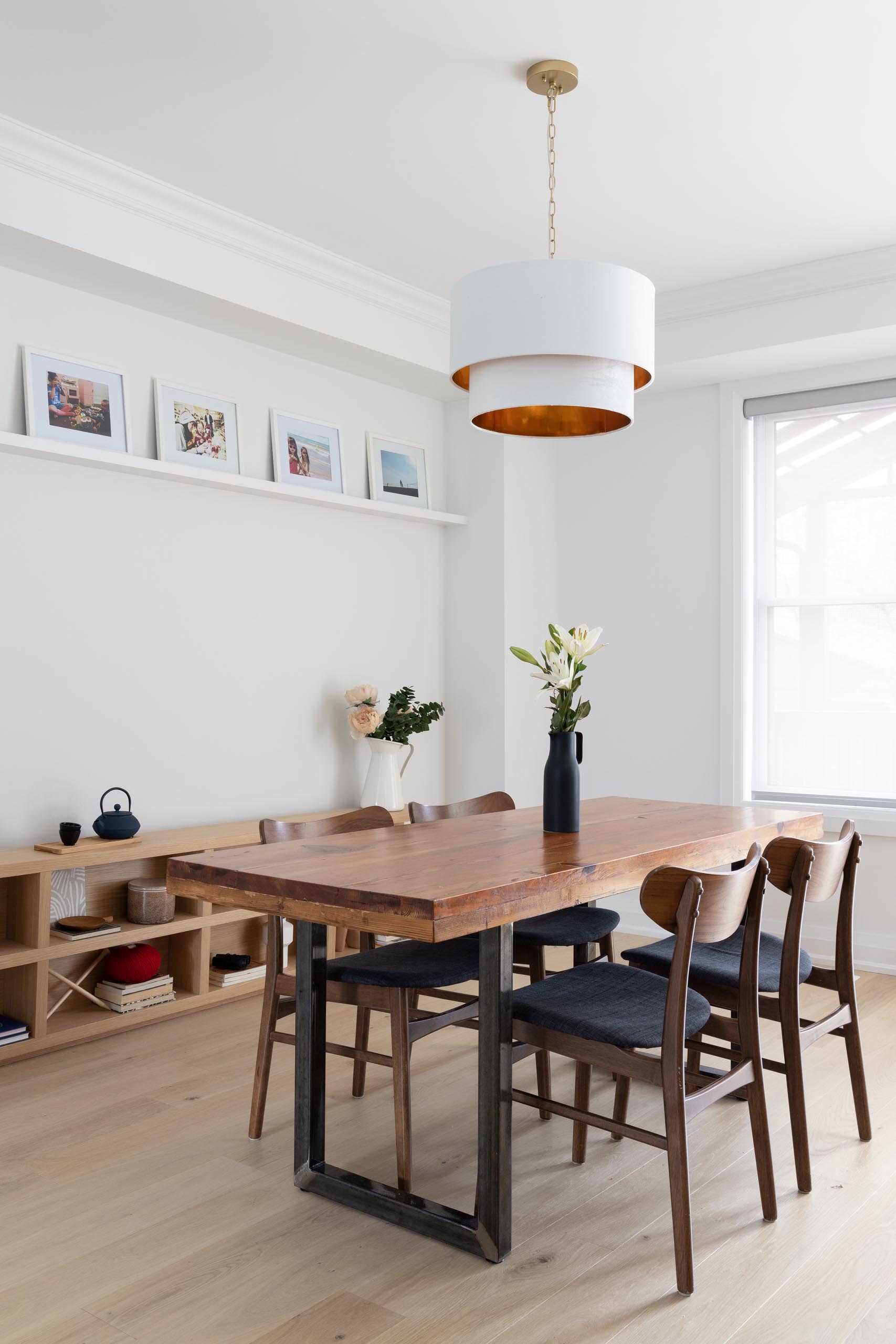 In this modern dining room, there's a wood table with a steel base that's located underneath a white pendant light with a metallic interior. A low wood console lines the wall, while a floating white shelf acts as a picture rail for family photos.