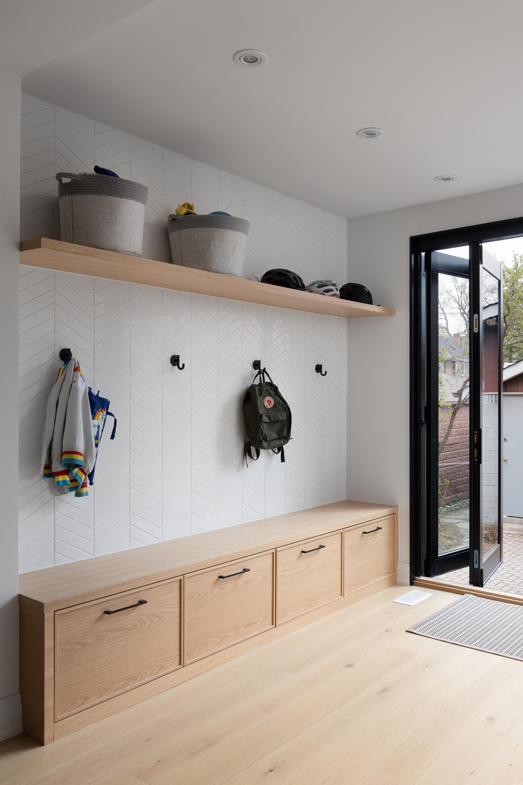 This mudroom includes a bench that includes storage, hooks for coats and bags, a floating wood shelf, and a chevron accent wall.