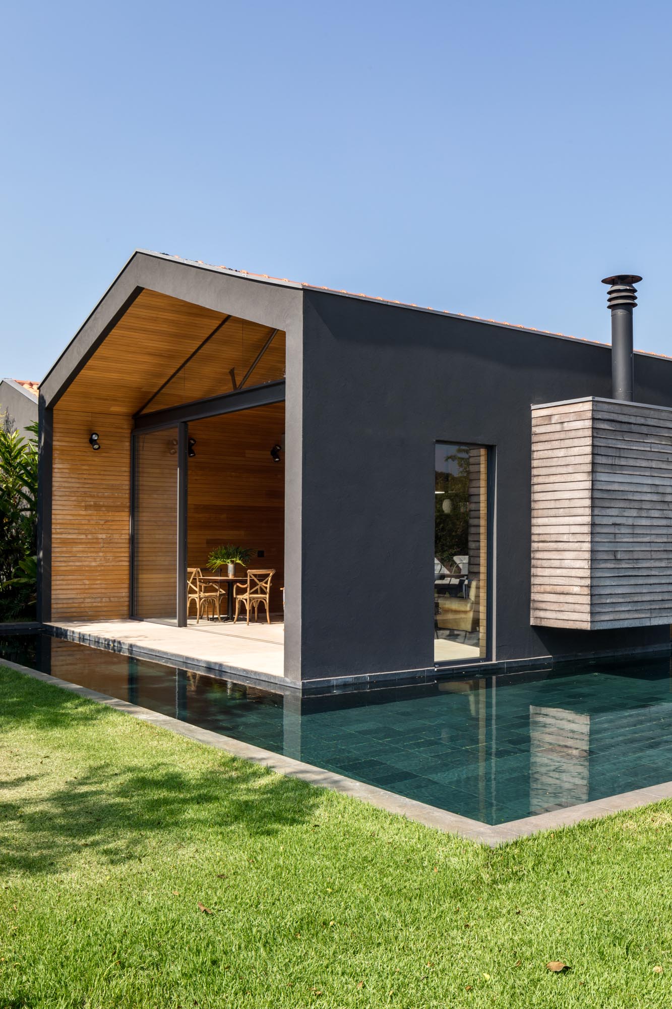 The exterior of this modern home is a black-painted mass, contrasting the surrounding nature, and providing a bold backdrop for the outdoors spaces, like the patio and swimming pool.