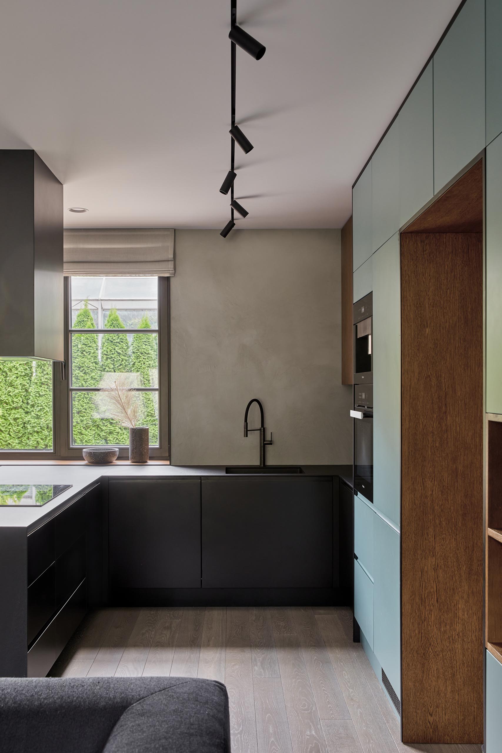 An open plan room living room, dining room, and kitchen, has a muted mint green wall that's filled with minimalist cabinetry and open wood shelving. The cabinets continue through the kitchen where they meet black cabinets and a matching countertop.