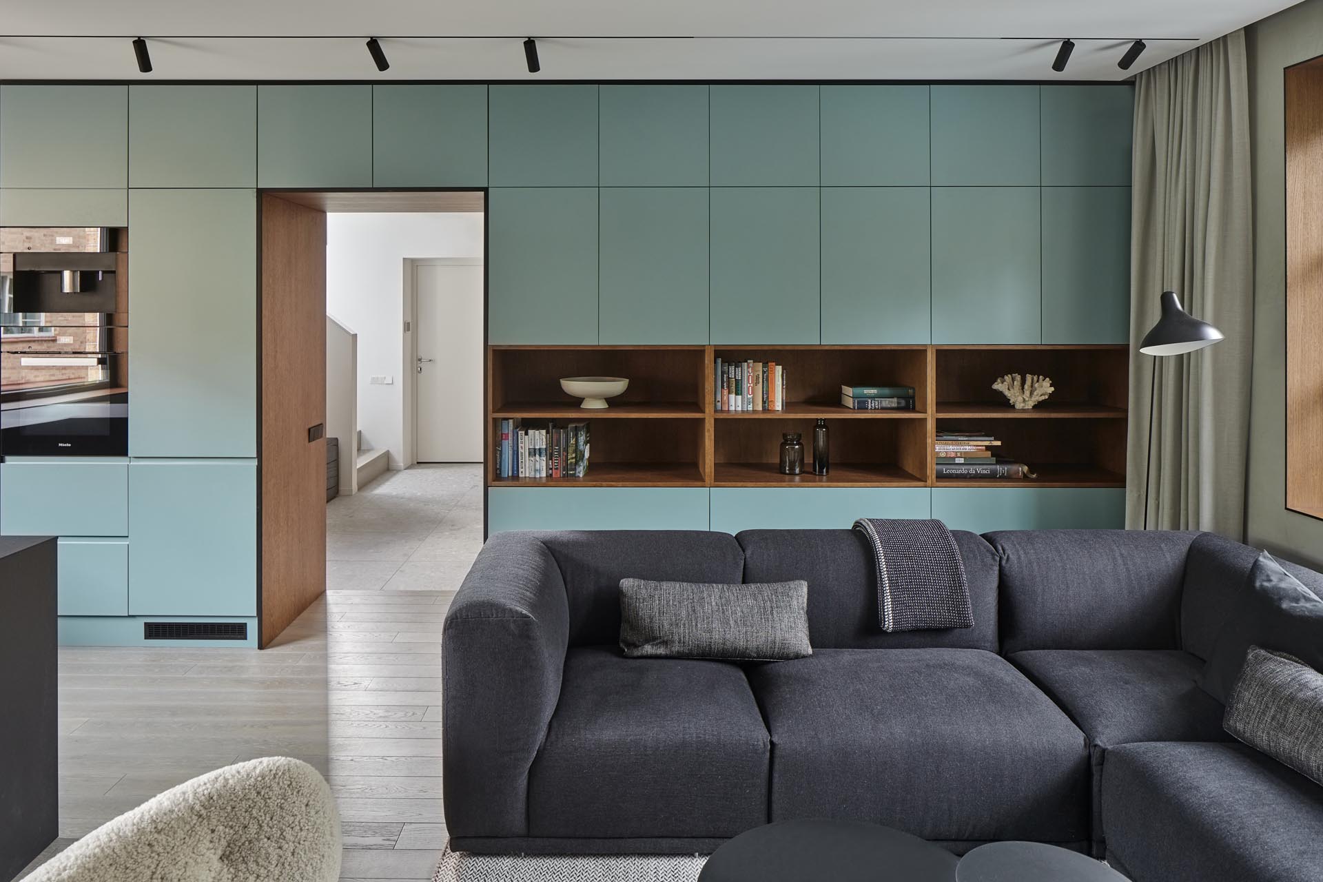 An open plan room living room, dining room, and kitchen, has a muted mint green wall that's filled with minimalist cabinetry and open wood shelving. The cabinets continue through the kitchen where they meet black cabinets and a matching countertop.