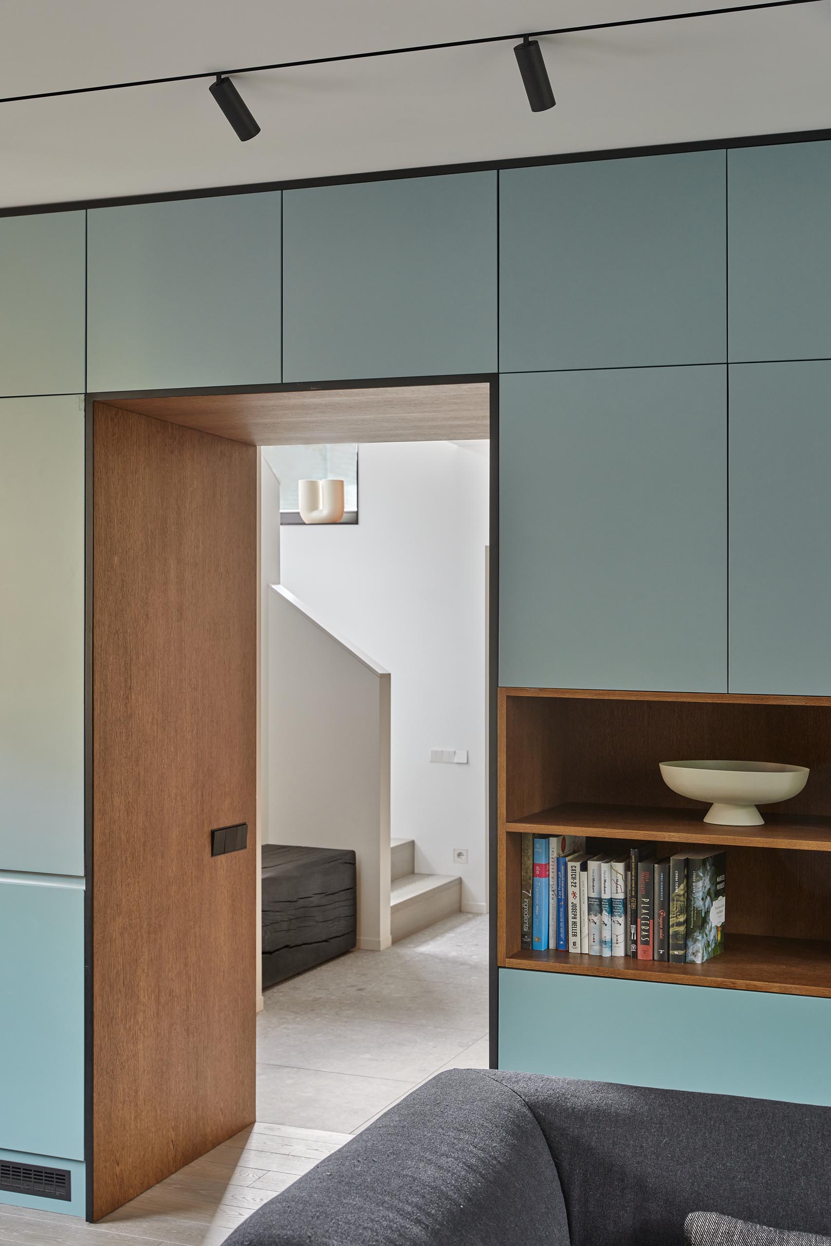 An open plan room living room, dining room, and kitchen, has a muted mint green wall that's filled with minimalist cabinetry and open wood shelving. The cabinets continue through the kitchen where they meet black cabinets and a matching countertop.