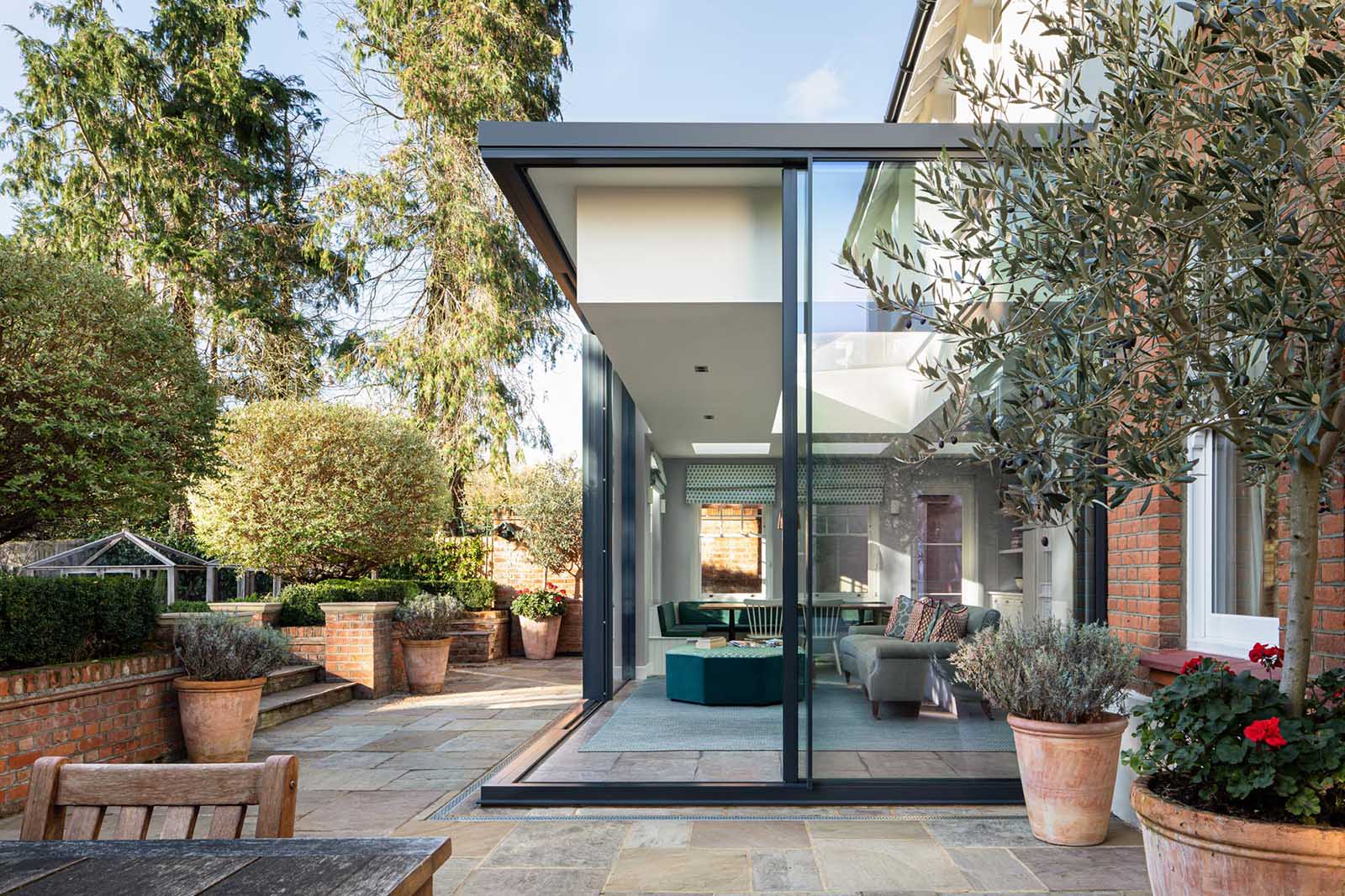 This new home addition, furnished with a dining area and living space, has a black frame with floor-to-ceiling walls of glass, which can be opened to connect the interior of the room with the outdoors.