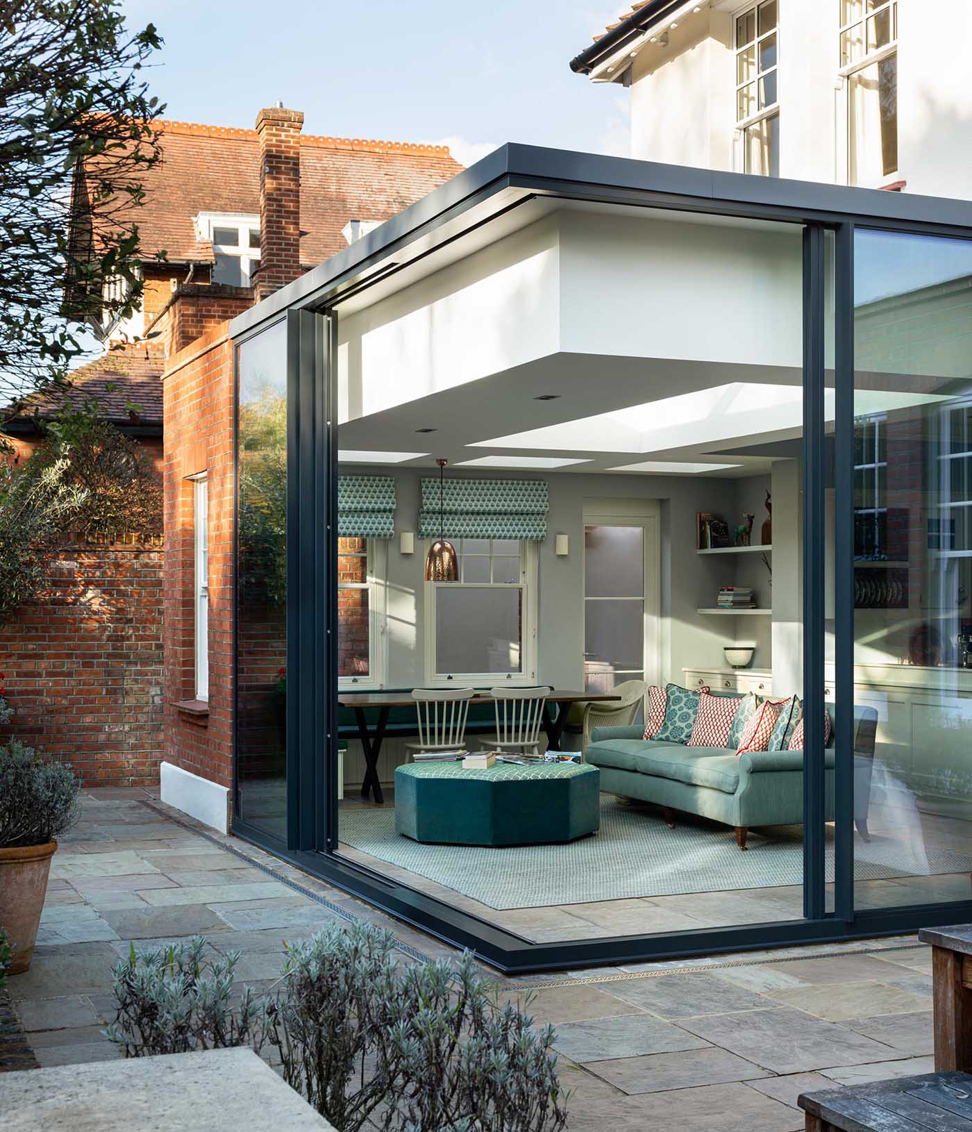 This new home addition, furnished with a dining area and living space, has a black frame with floor-to-ceiling walls of glass, which can be opened to connect the interior of the room with the outdoors.