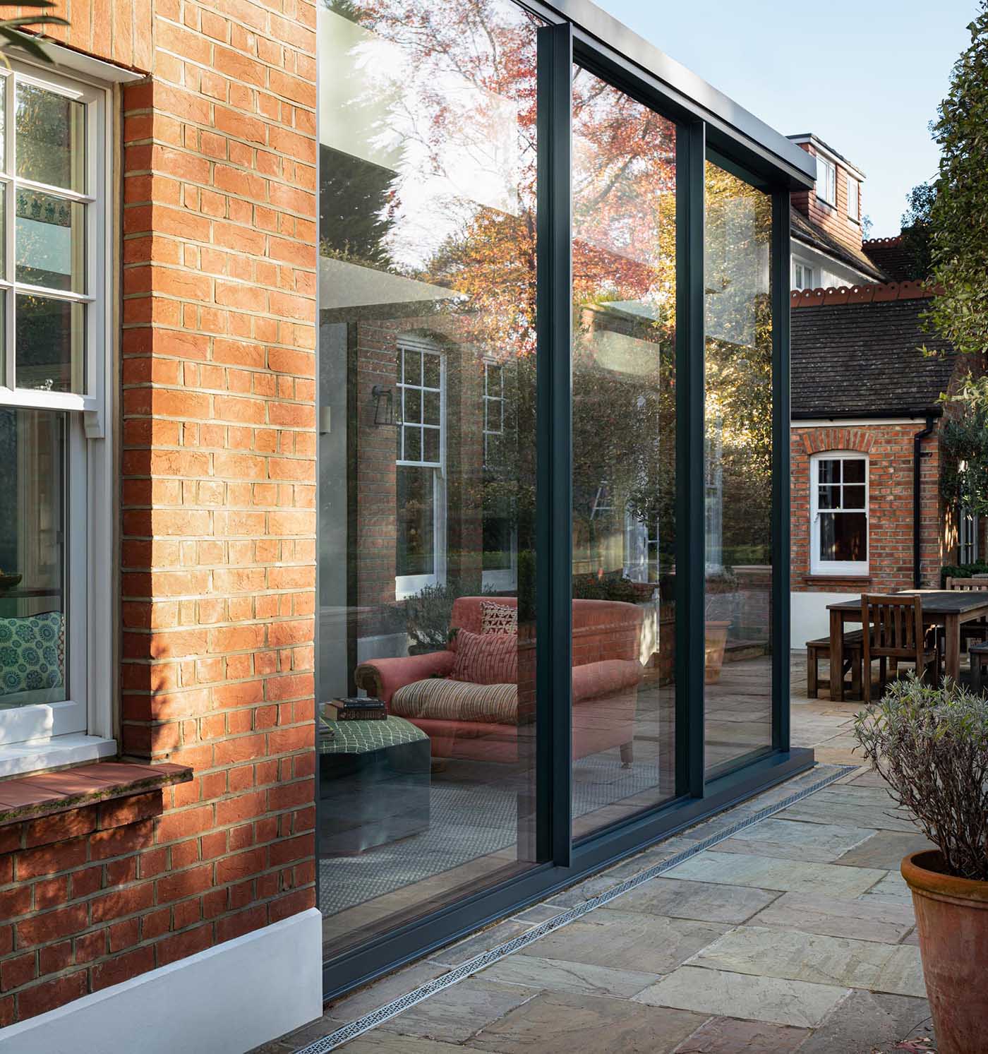 This new home addition, furnished with a dining area and living space, has a black frame with floor-to-ceiling walls of glass, which can be opened to connect the interior of the room with the outdoors.