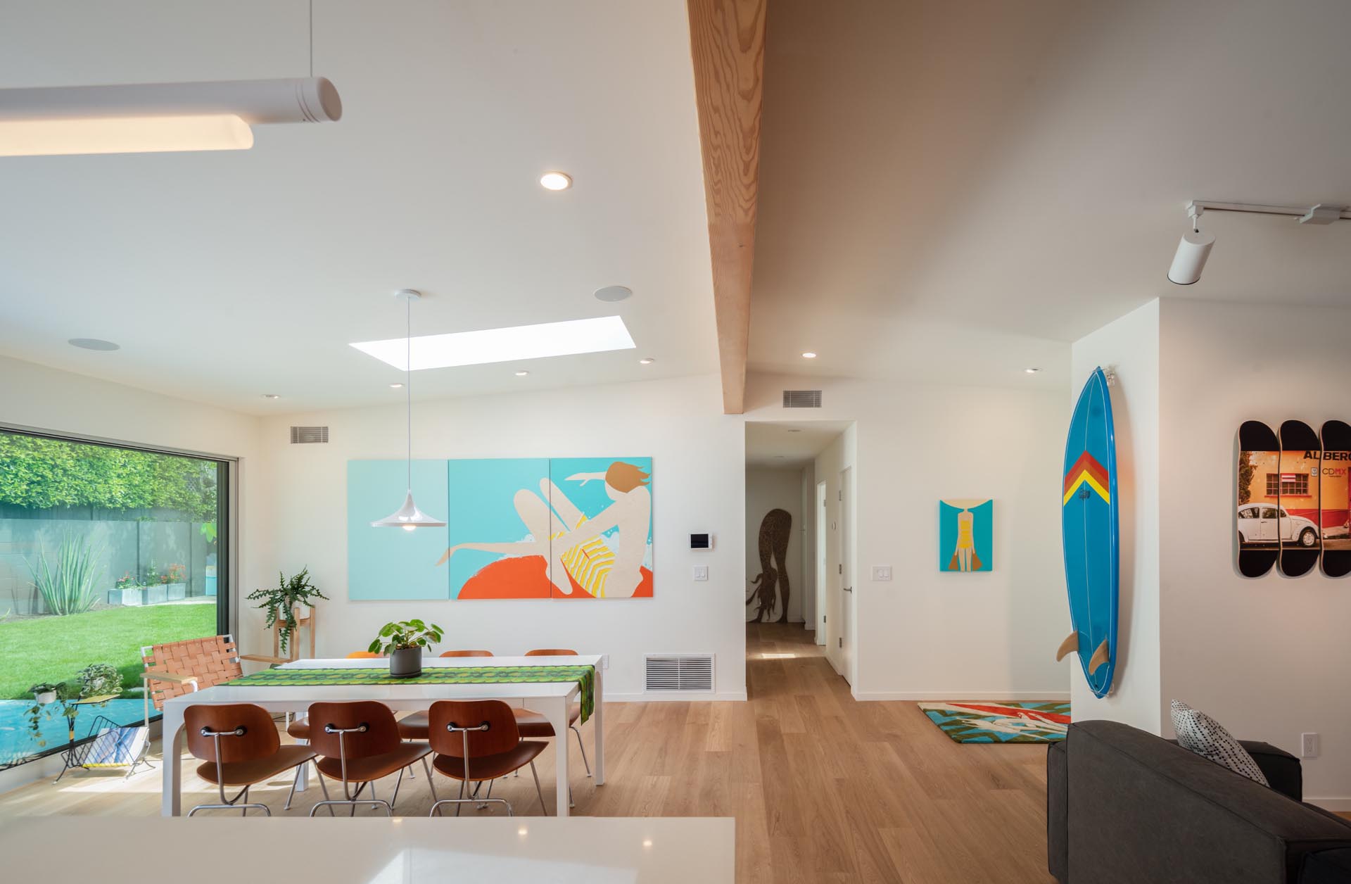 This modern open plan dining area has been furnished with a white dining table and a colorful 3-panel art piece on the wall. A skylight adds natural light to the space.