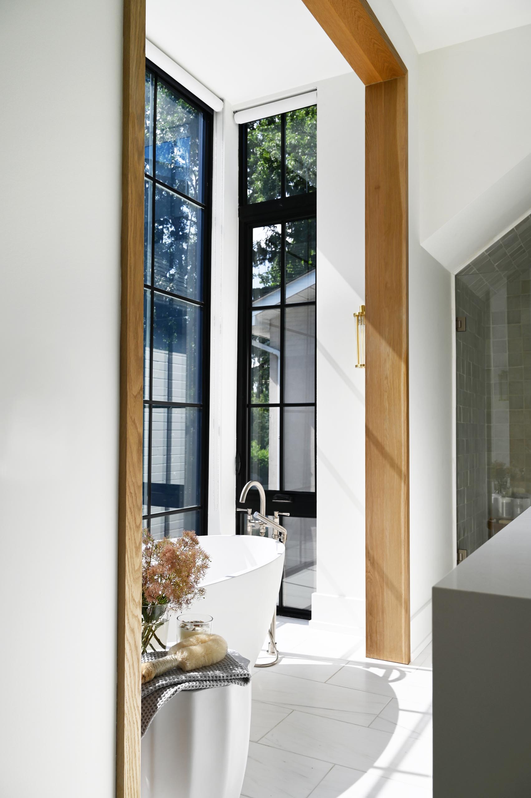 This modern white marbled spa-like bathroom includes a windowed alcove for the freestanding tub, metallic hardware, and wood accents.