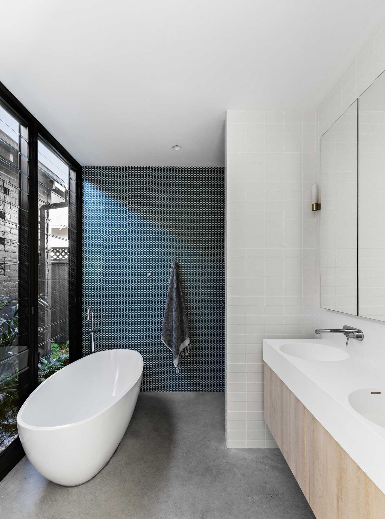 A modern bathroom with a green penny tile accent wall, freestanding bathtub, louvre windows, and concrete flooring.