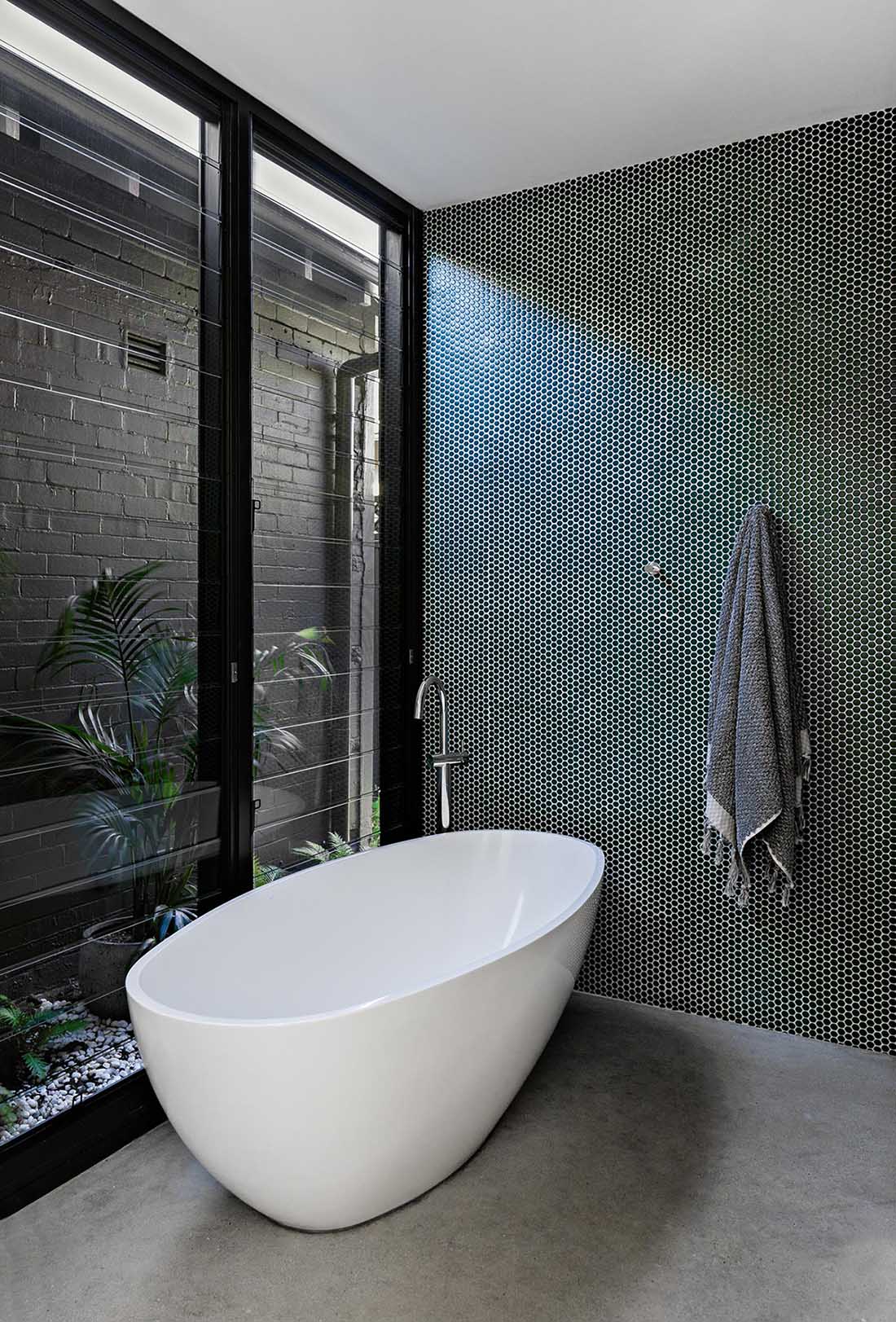 A modern bathroom with a green penny tile accent wall, freestanding bathtub, louvre windows, and concrete flooring.