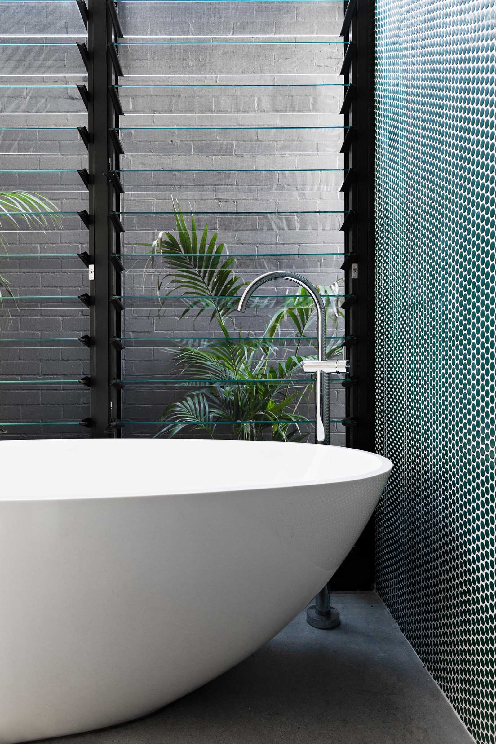 A modern bathroom with green penny tile accent wall, freestanding bathtub, and louvre windows.