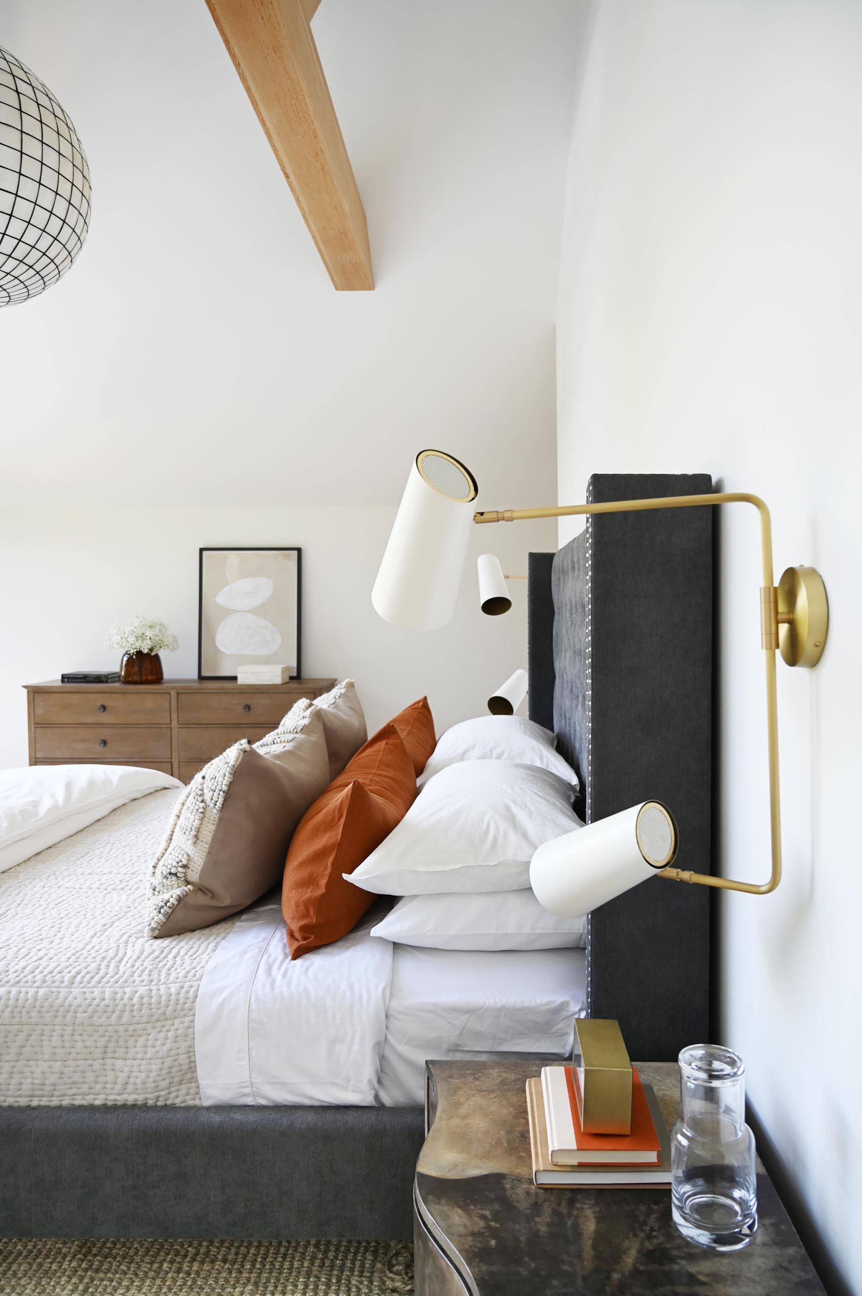 Featuring a cathedral ceiling with exposed beams and layers of neutral soft textures and furnishings, this primary bedroom is designed as a cozy nest overlooking the lush green yard.